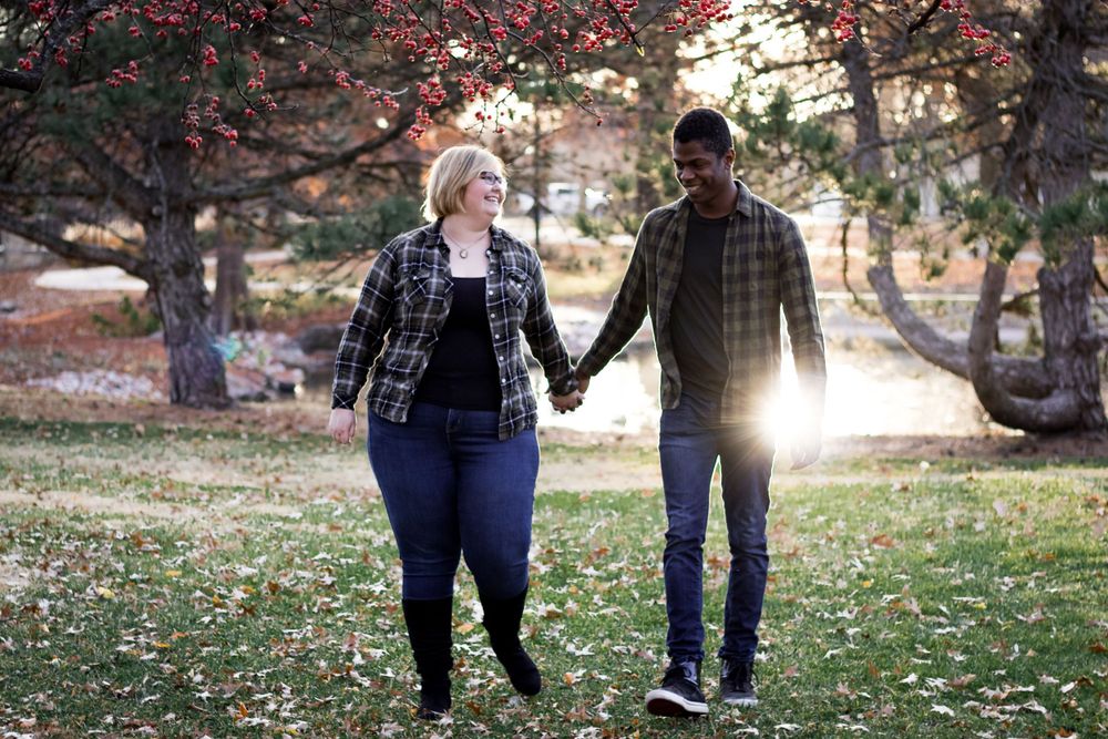 A couple walks hand in hand through a sunlit autumn park with fallen leaves on the ground and trees overhead.