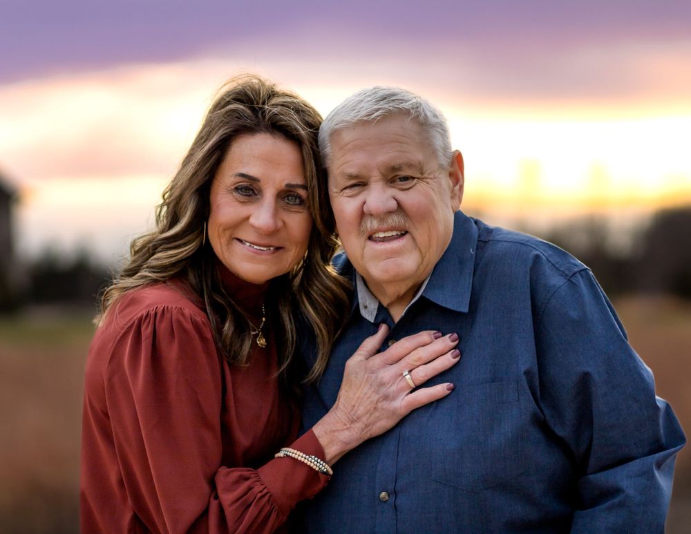 Two people wearing red and blue jackets embrace while posing together outdoors at sunset.