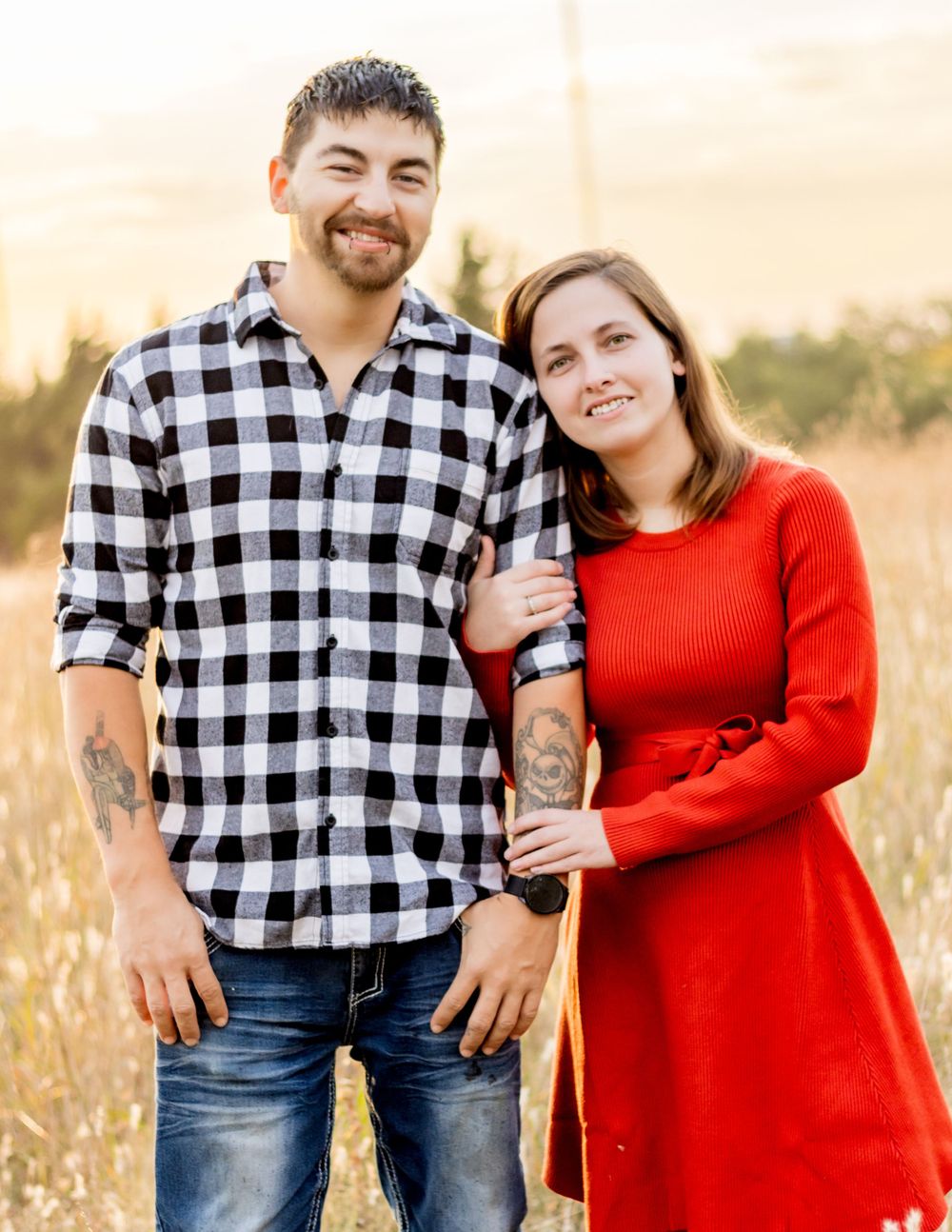 A couple stands together in a golden field at sunset, wearing a black and white plaid shirt and red dress.