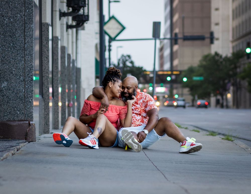 Two figures sit together on a city sidewalk wearing bright pink outfits and sneakers on a summer evening.