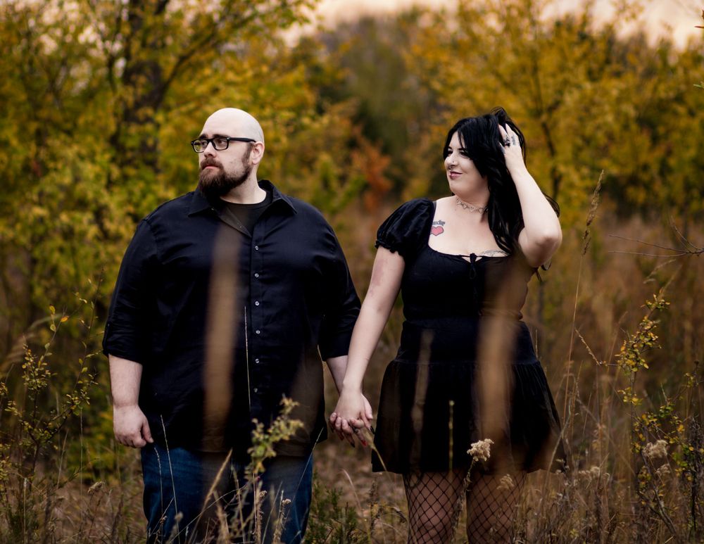 A couple in matching black clothing holds hands while standing in a field during autumn.