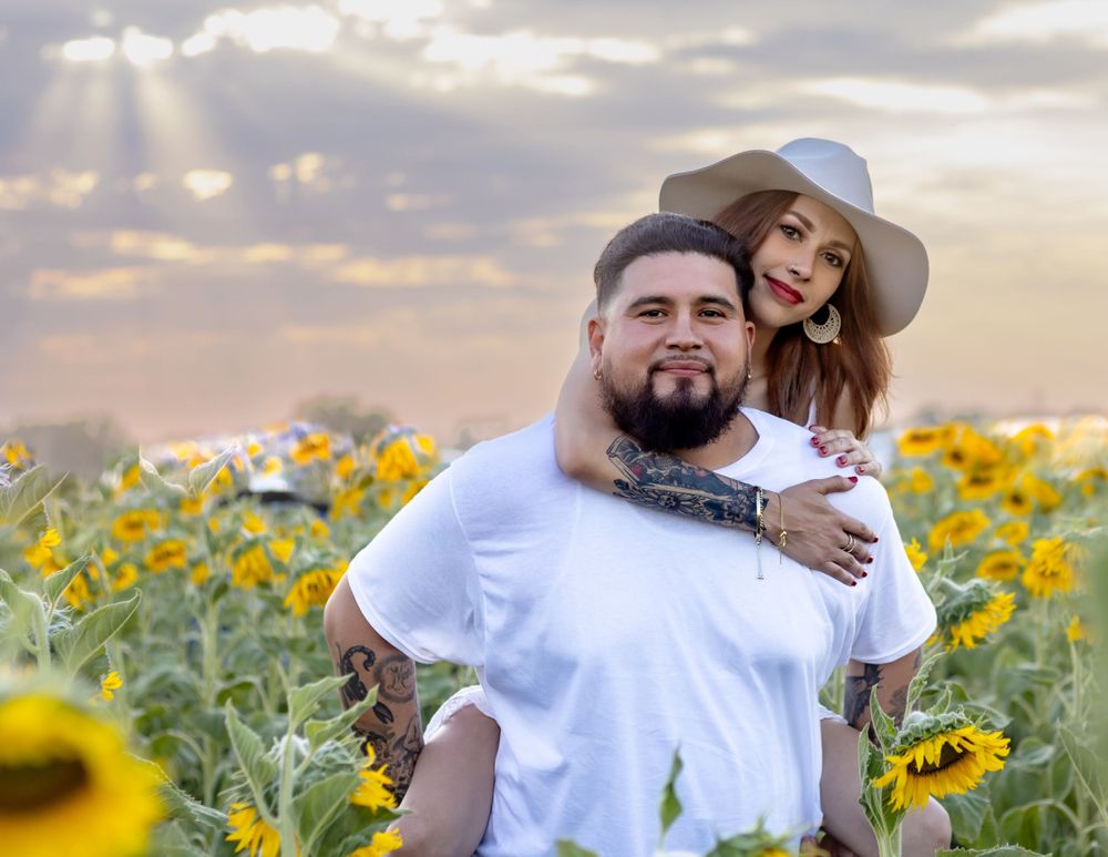 Two people embracing in a sunflower field at sunset with sunbeams peeking through clouds in the background.