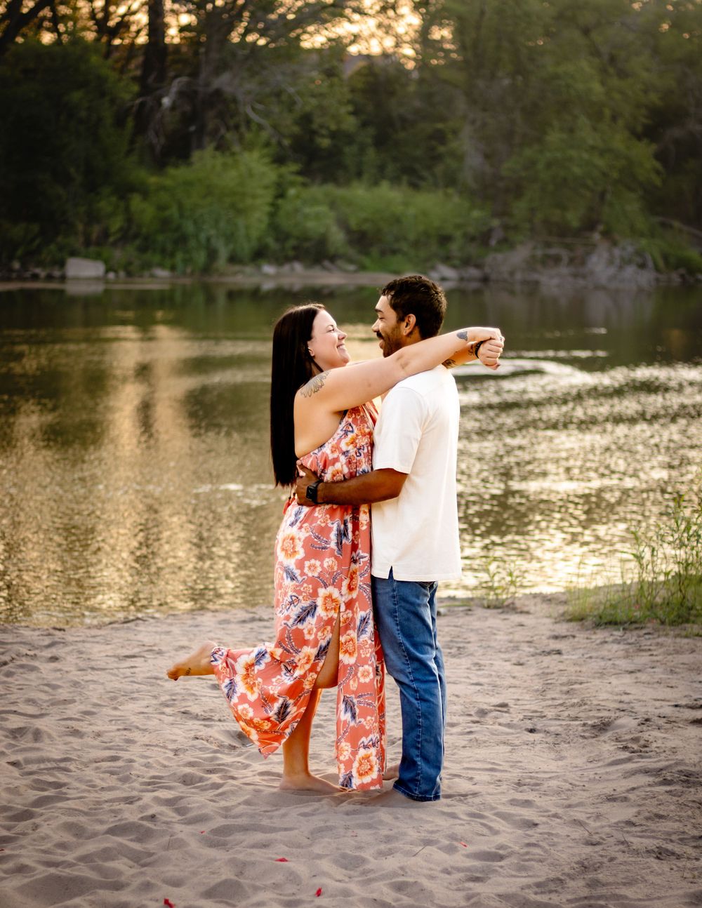 A couple embracing by a tranquil lake at sunset, with the woman wearing a flowing floral dress while standing on the beach.