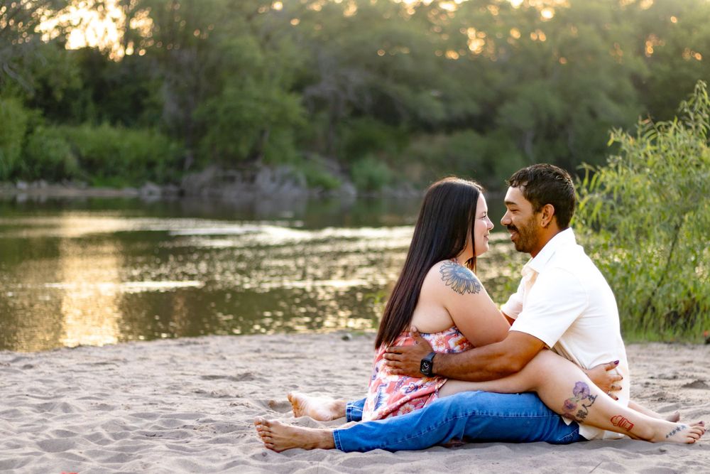 Couple sitting together on sandy riverbank at sunset with water and greenery in background.