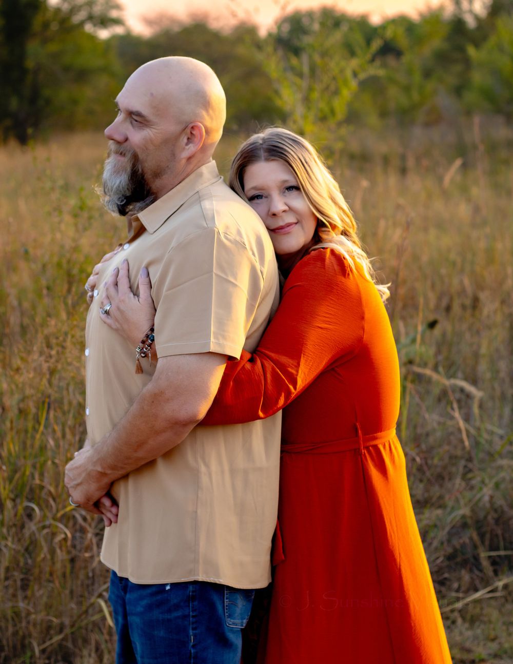 A couple embraces during a sunset photoshoot in a tall grass field, with warm golden light illuminating the scene.