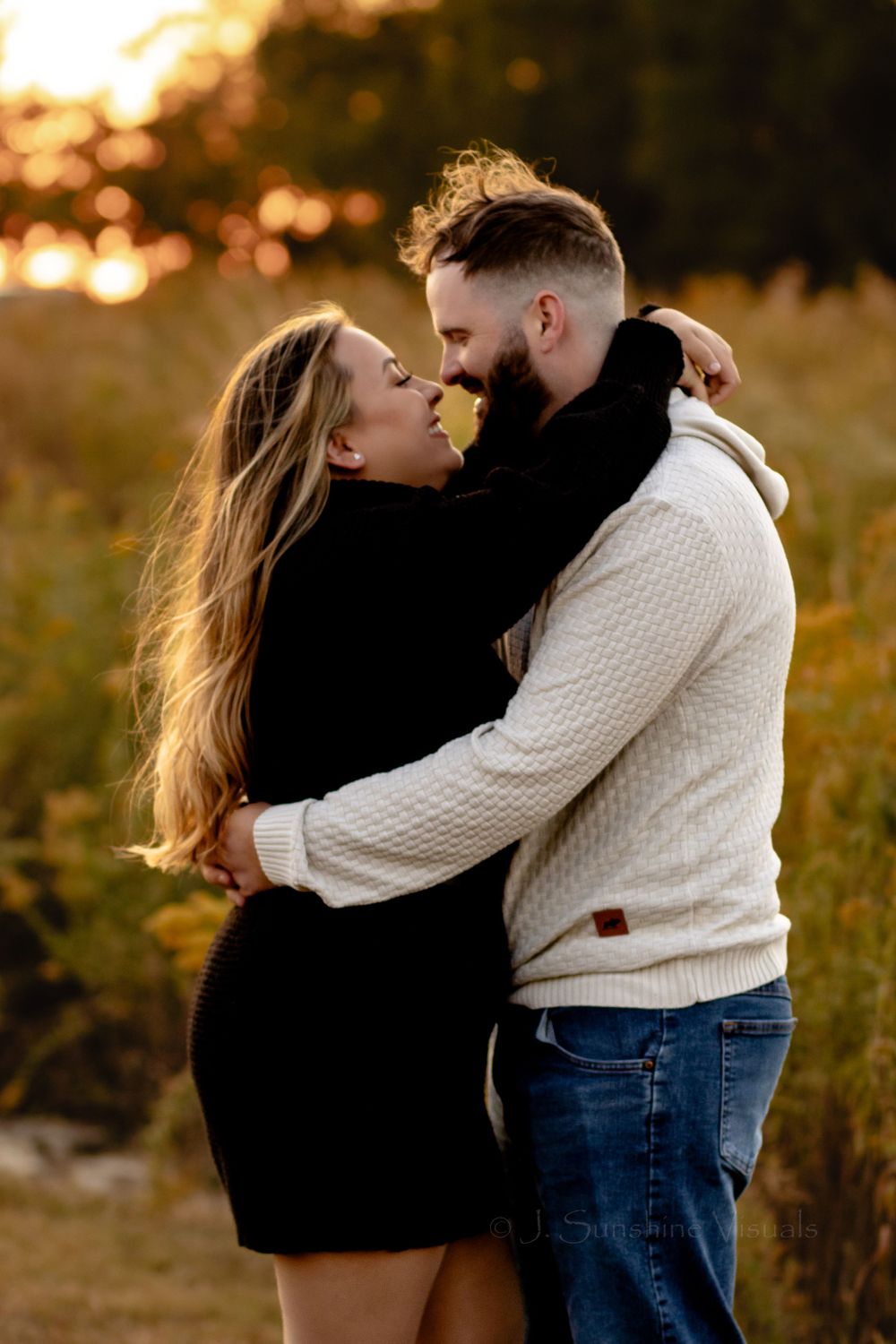 Romantic couple embraces during golden hour sunset, with woman in black dress and man wearing cream cable knit sweater.