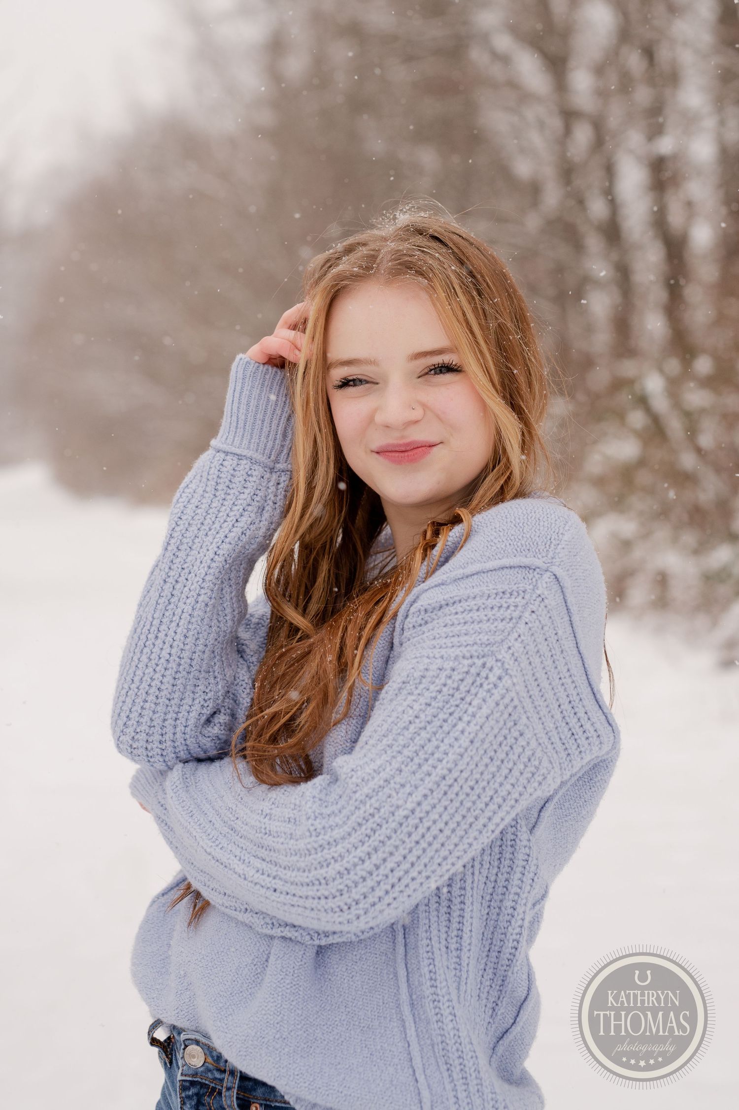 A winter portrait showing someone in a cozy sweater smiling against snowy trees.