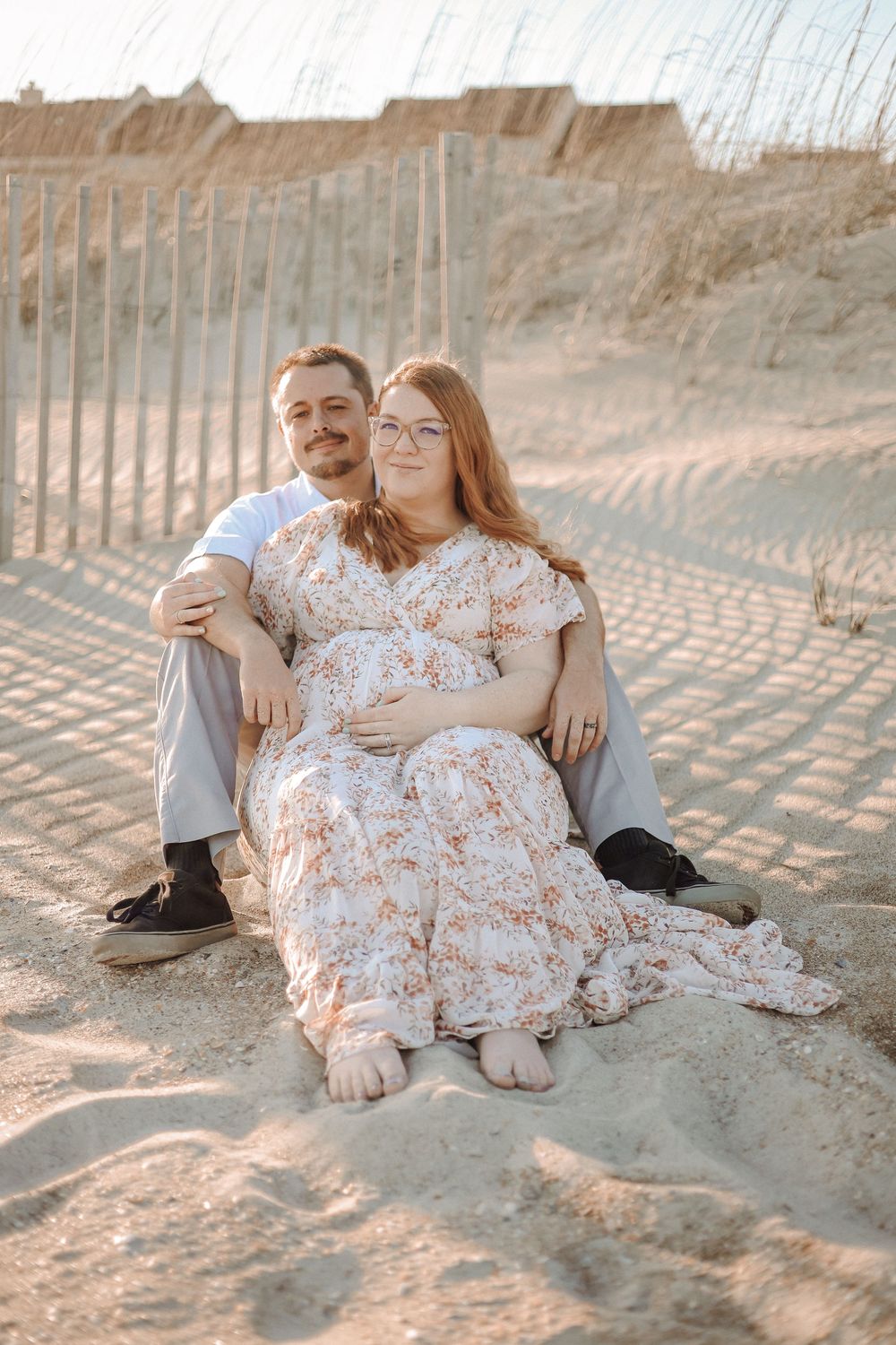 Coastal maternity photographer in NC — couple posing on a beach walkway surrounded by dunes during sunset.