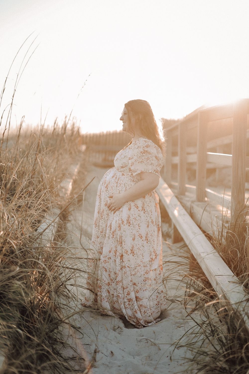Beach maternity photographer in Eastern North Carolina — pregnant woman in a white dress walking along a coastal boardwalk at sunset.
