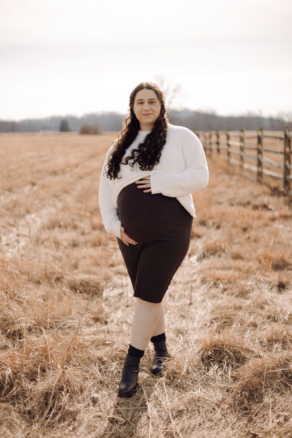 Winter maternity portrait in white sweater against rustic fence backdrop.