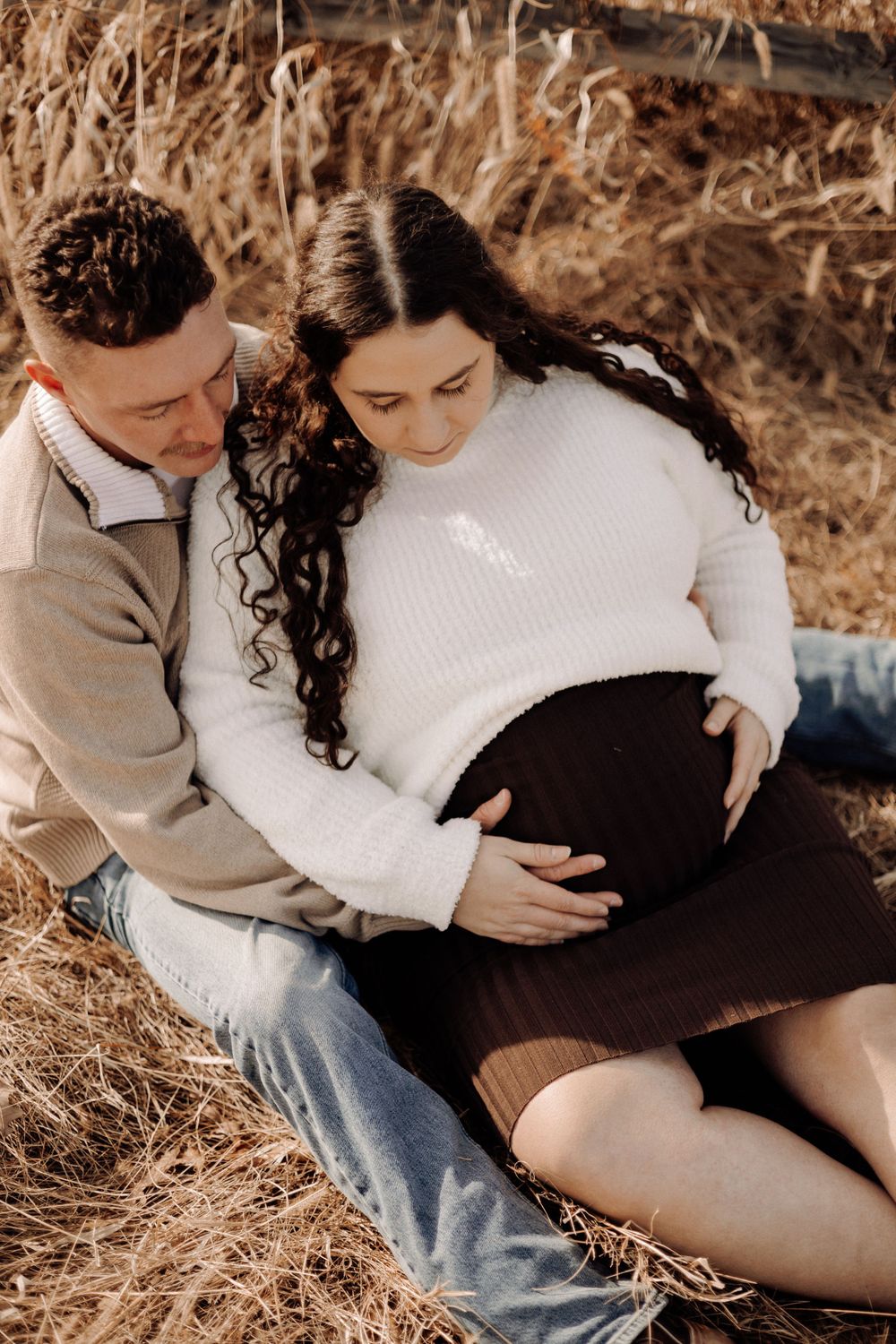 Expecting couple embraces tenderly while sitting in a golden field during sunset.