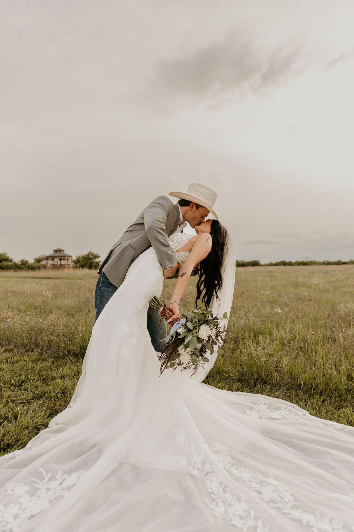 A couple shares a romantic dip kiss in an open field, with the bride wearing a flowing white dress with long train.