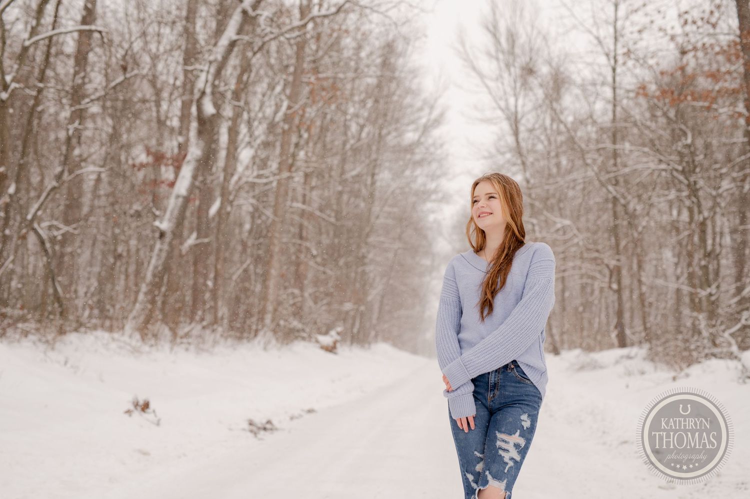 A person in a gray sweater and distressed jeans poses playfully in a winter forest setting.