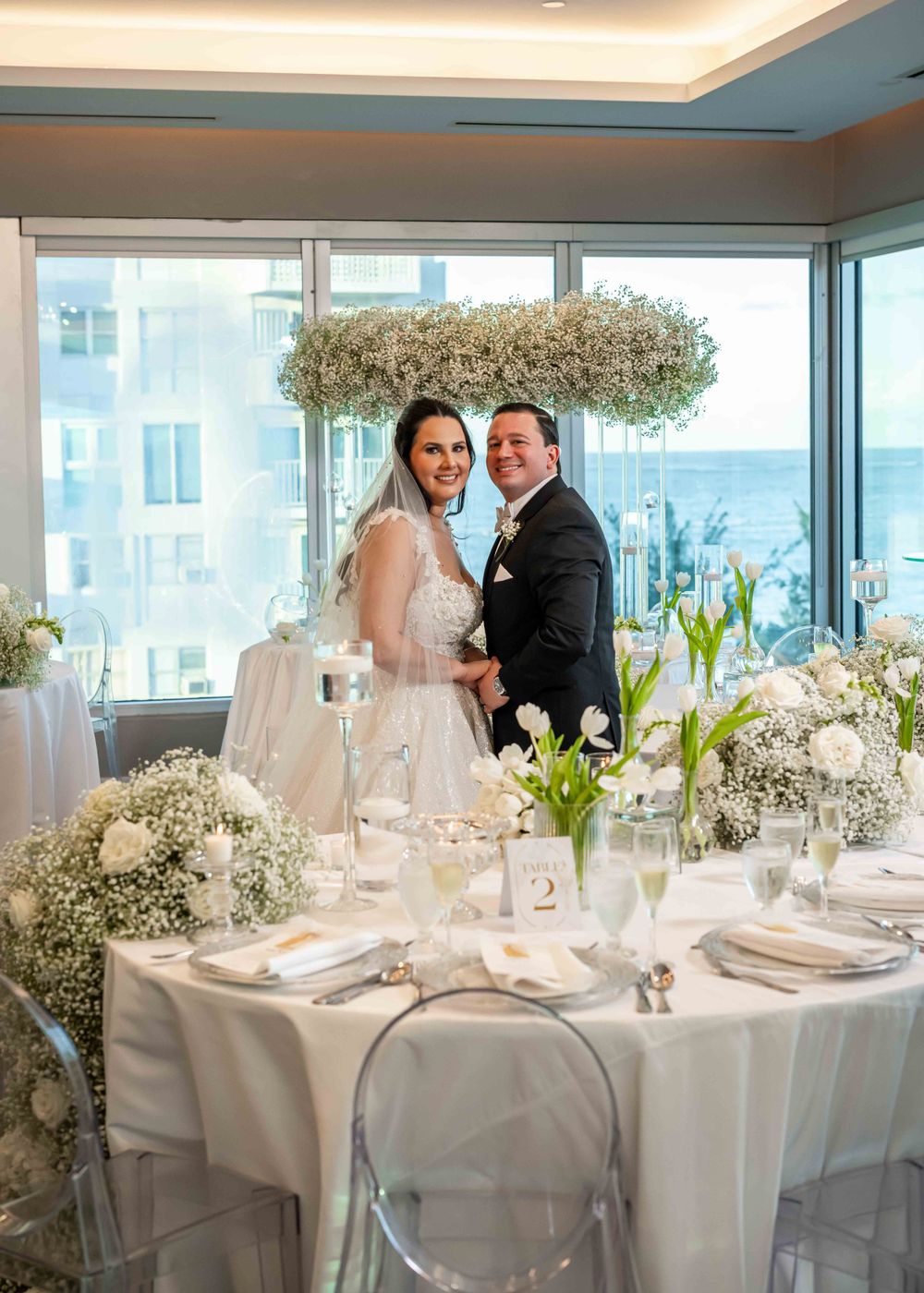 A wedding reception in a bright room with white table settings, baby's breath floral arrangements and panoramic windows.