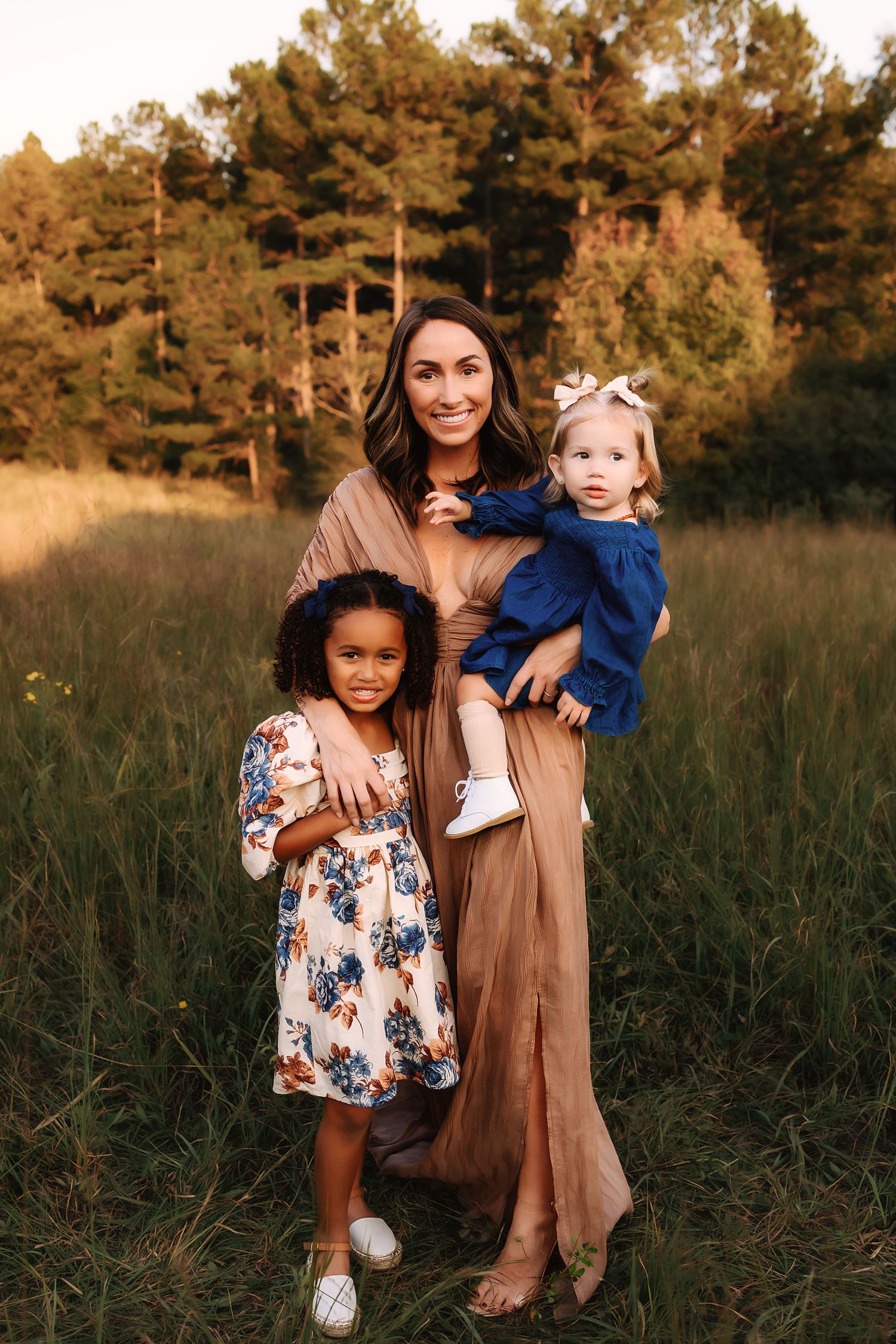 A family poses together outdoors during autumn with fall foliage and golden sunlight in the background.