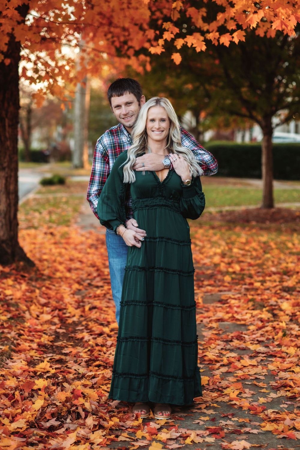 A couple embraces among fallen autumn leaves beneath trees with vibrant orange foliage in a park setting.