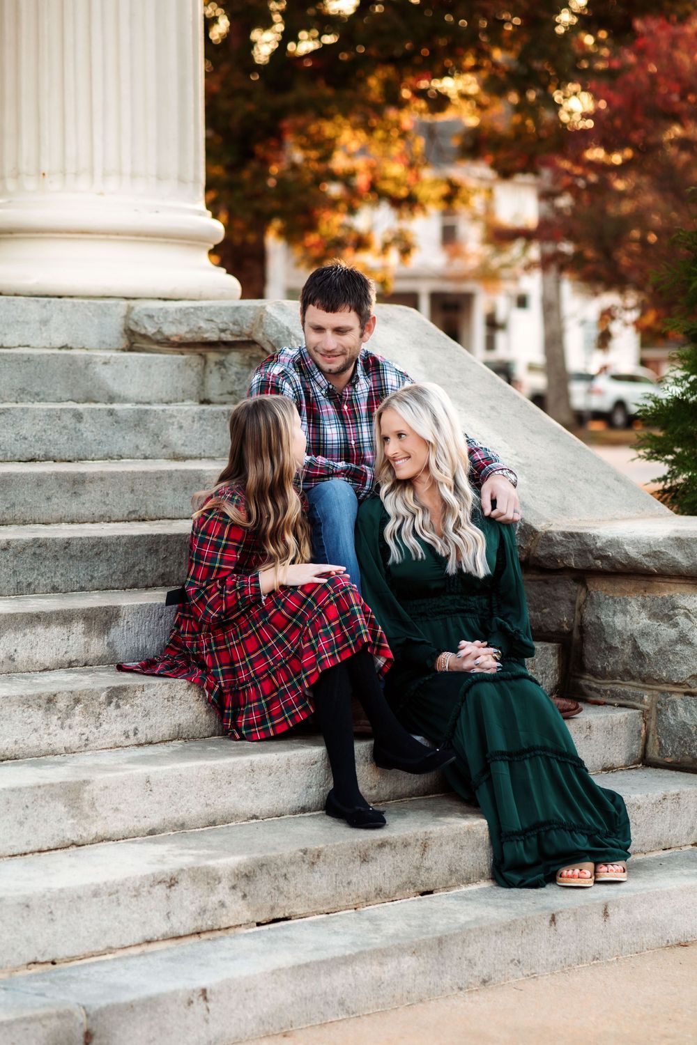 Three people sitting on stone steps wearing plaid and green dresses during autumn with columns and foliage in background.