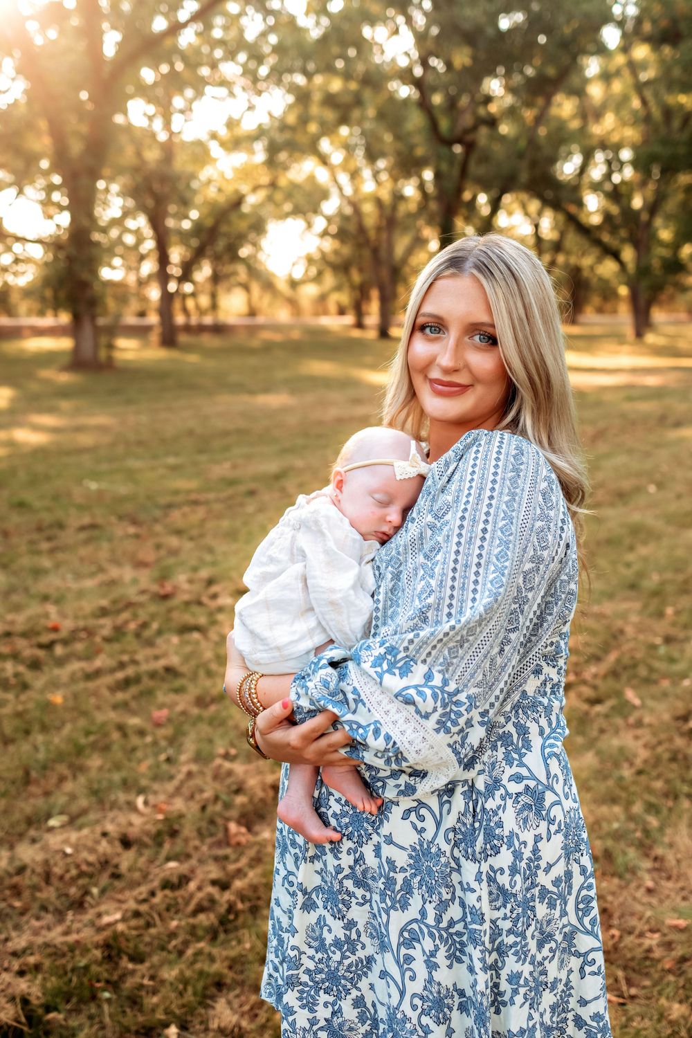A person in a blue and white floral dress holds a baby in white clothing during golden hour in a peaceful park setting.