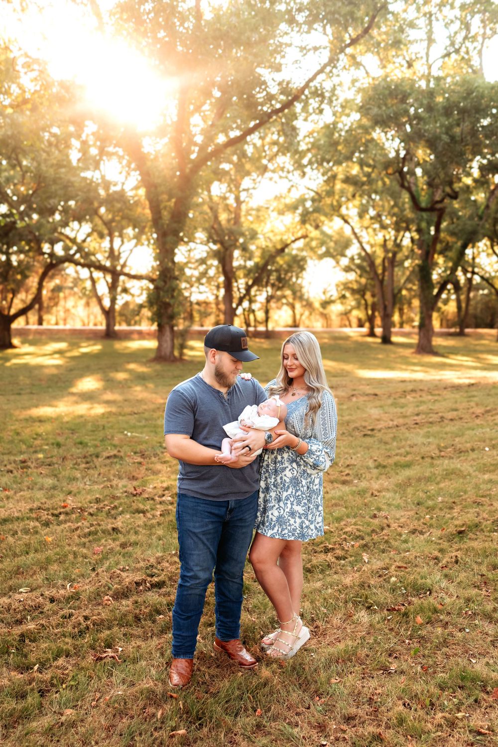 A couple holds their baby outdoors in a park during golden hour sunset with oak trees in the background.
