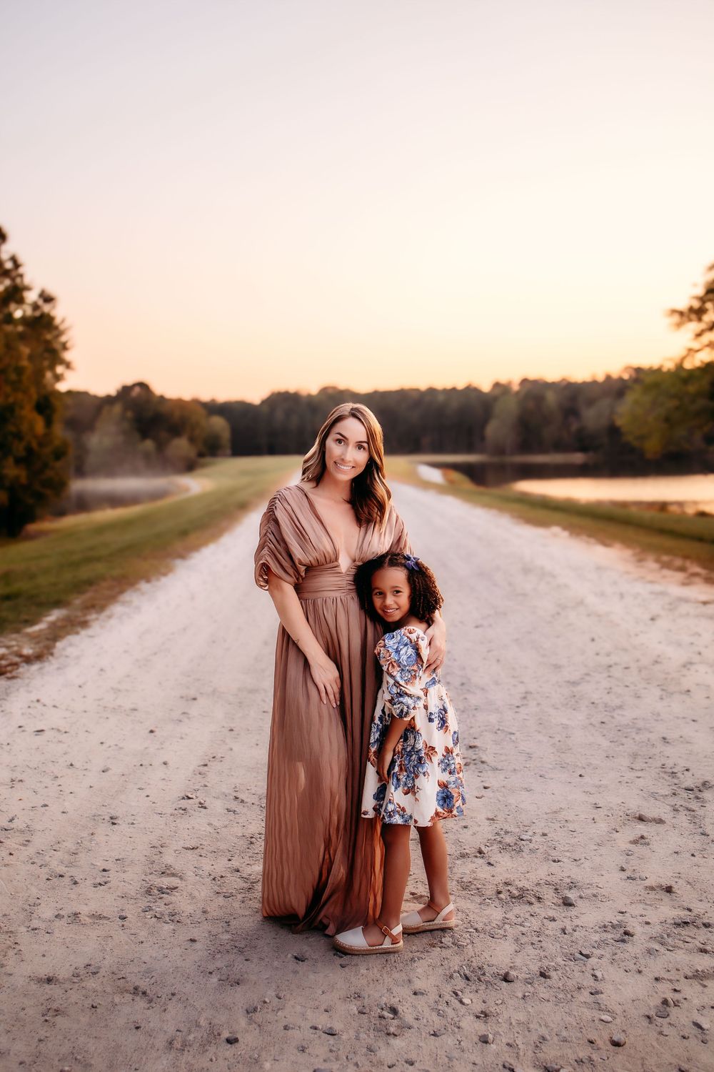 Two people in elegant dresses stand together on a dirt path surrounded by trees at sunset.