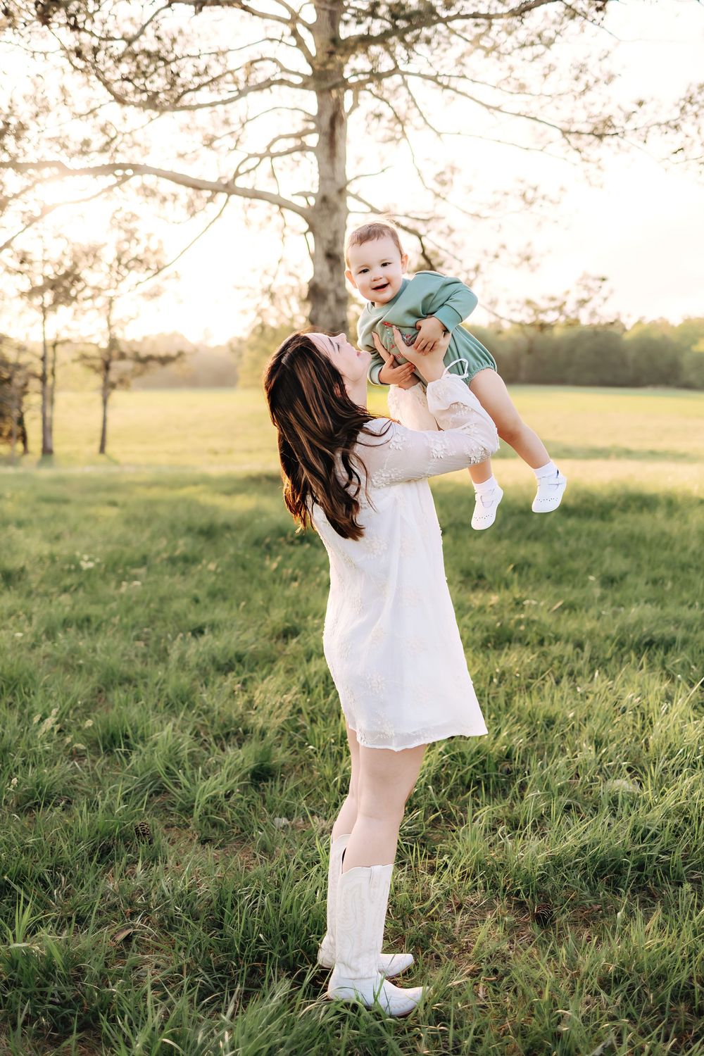 A woman in a white dress lifts a small child joyfully in a sunlit meadow at golden hour.