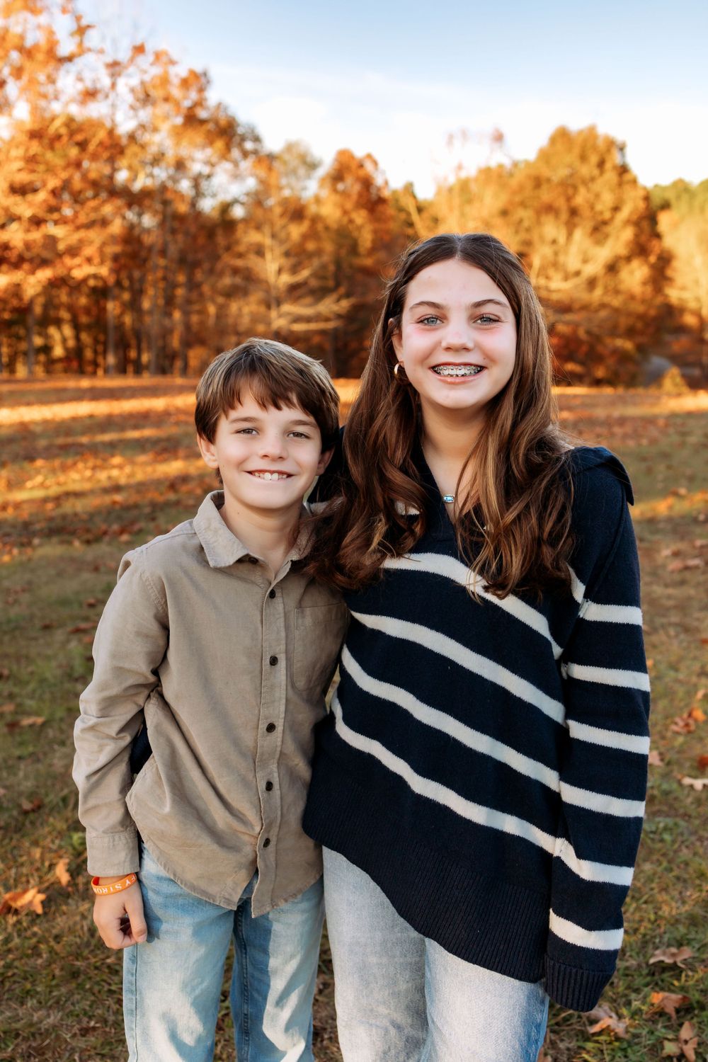 Two children stand together smiling outdoors on an autumn day with golden fall foliage in the background.
