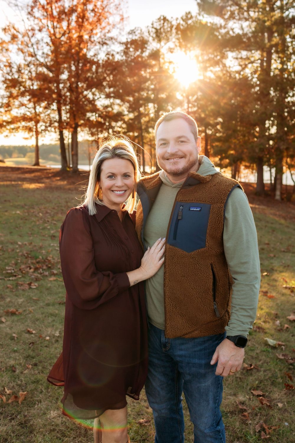 A couple stands together outdoors during golden hour sunset with fall trees and warm lighting in the background.