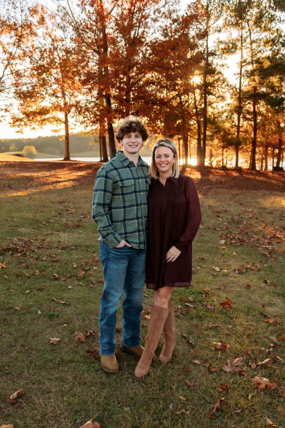 A couple stands together on an autumn hillside at sunset with golden light filtering through trees behind them.