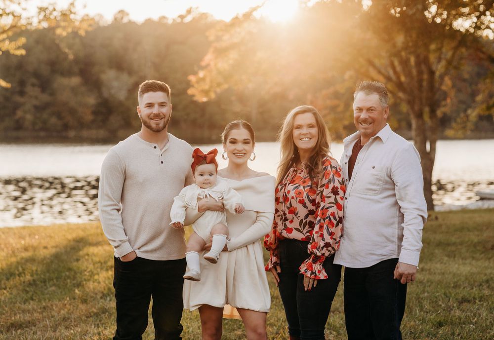 A family poses together by a serene lake at sunset with golden sunlight filtering through trees in the background.