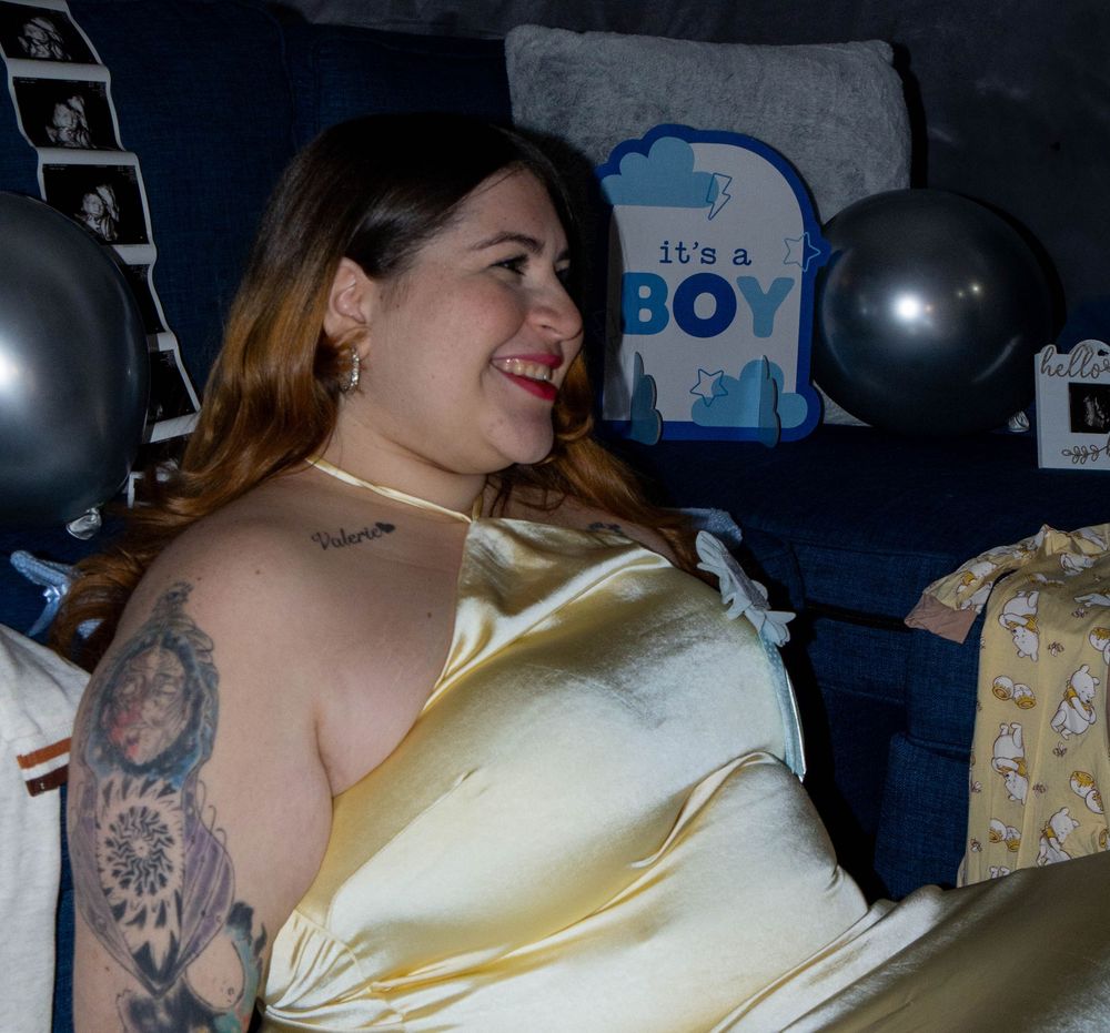 A person in a shiny gold dress sits and smiles while showing tattoos on their arm in a dimly lit room.