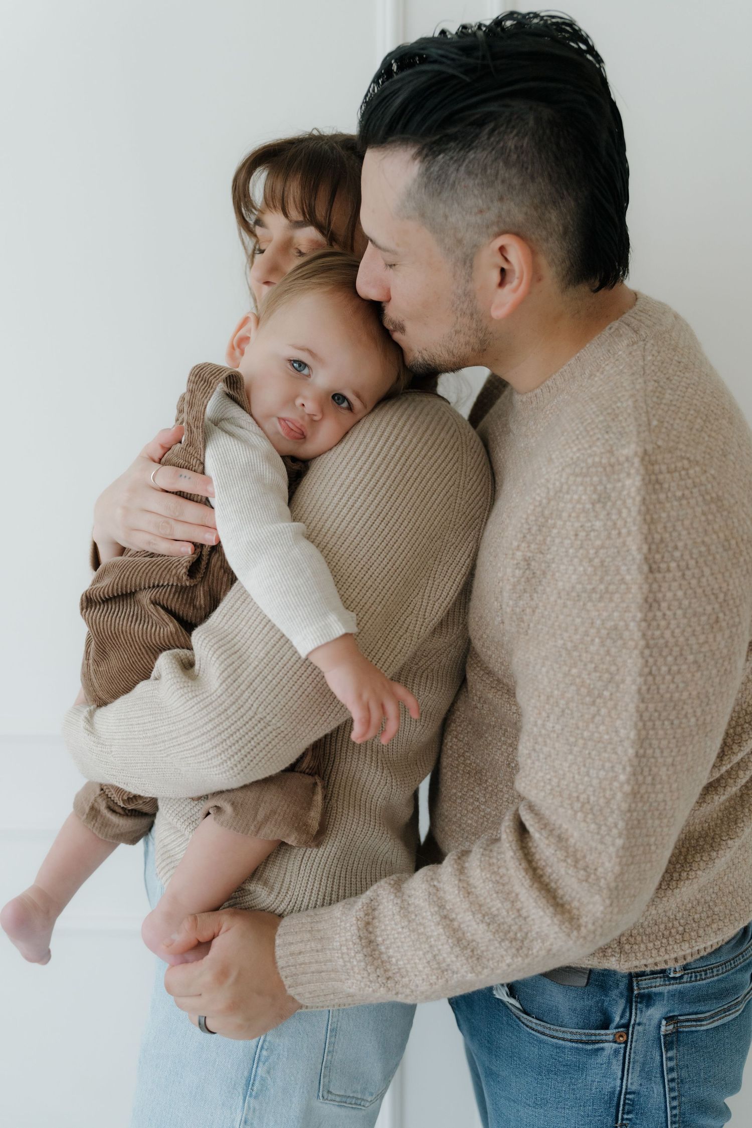 A tender family moment captured in neutral tones as they embrace their baby against a white wall.