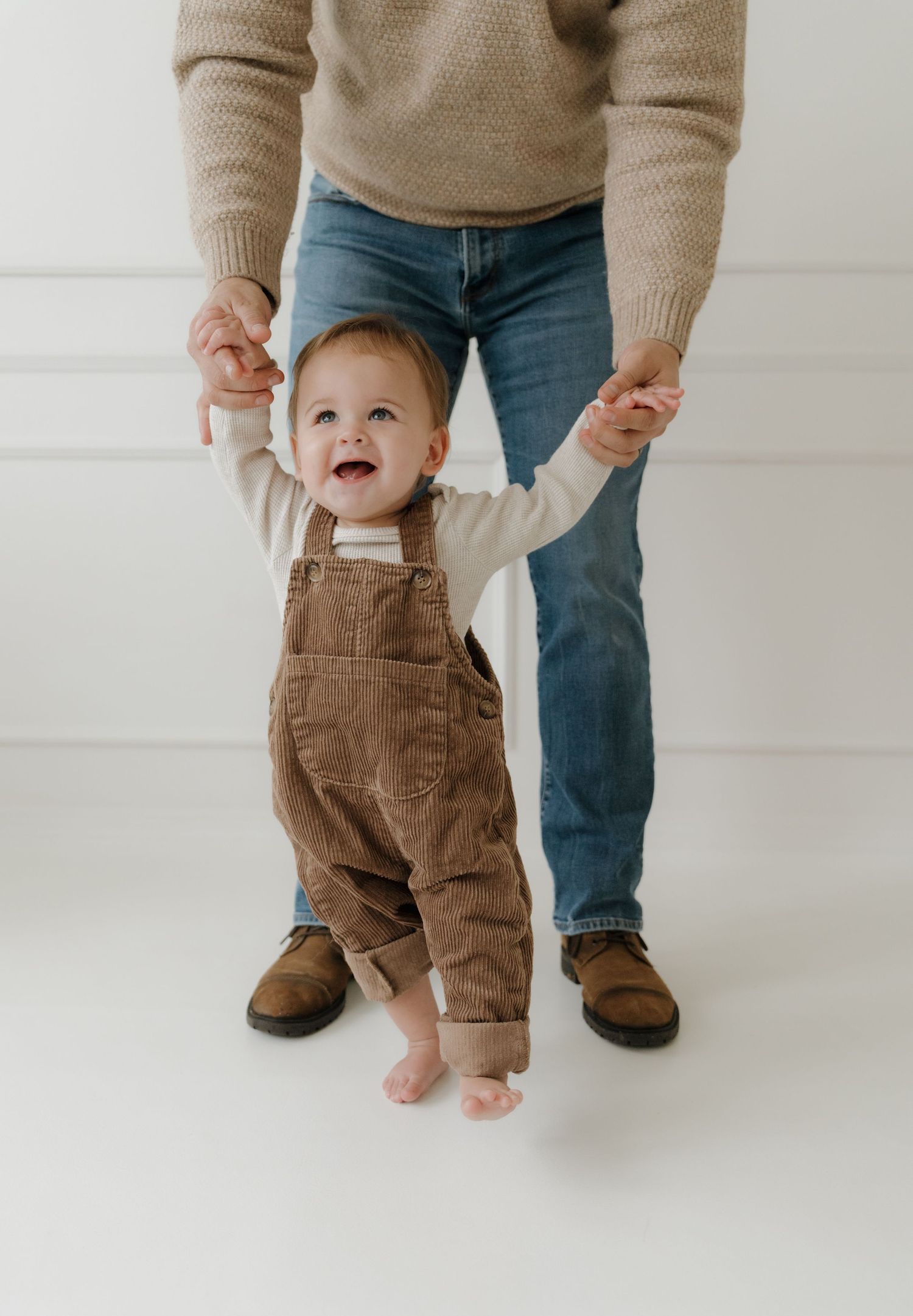 A parent holds a baby wearing brown corduroy pants while standing against a white wall in a progression of playful poses.