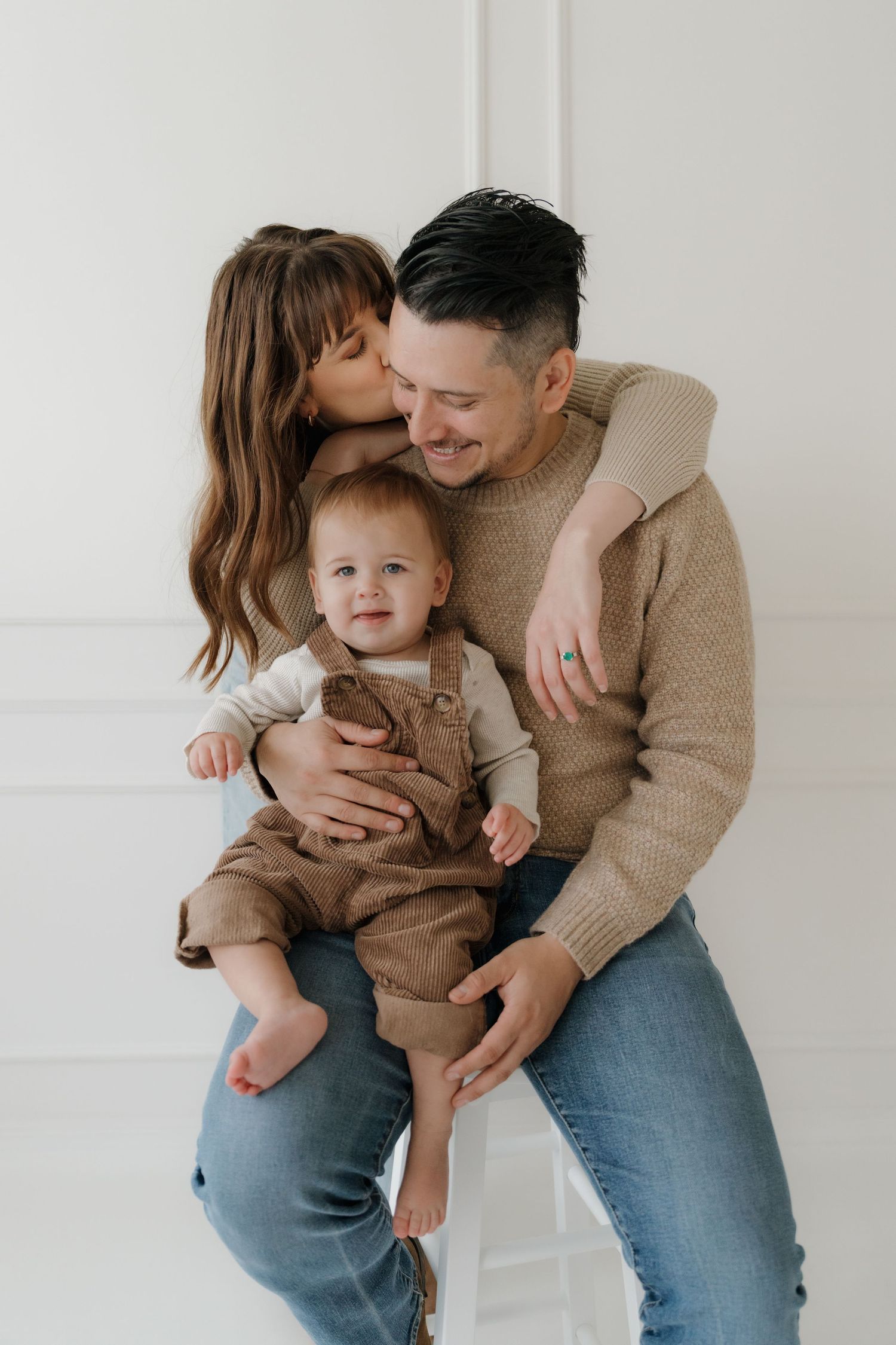 Intimate family portrait in matching beige sweaters sharing loving moments against a white backdrop.