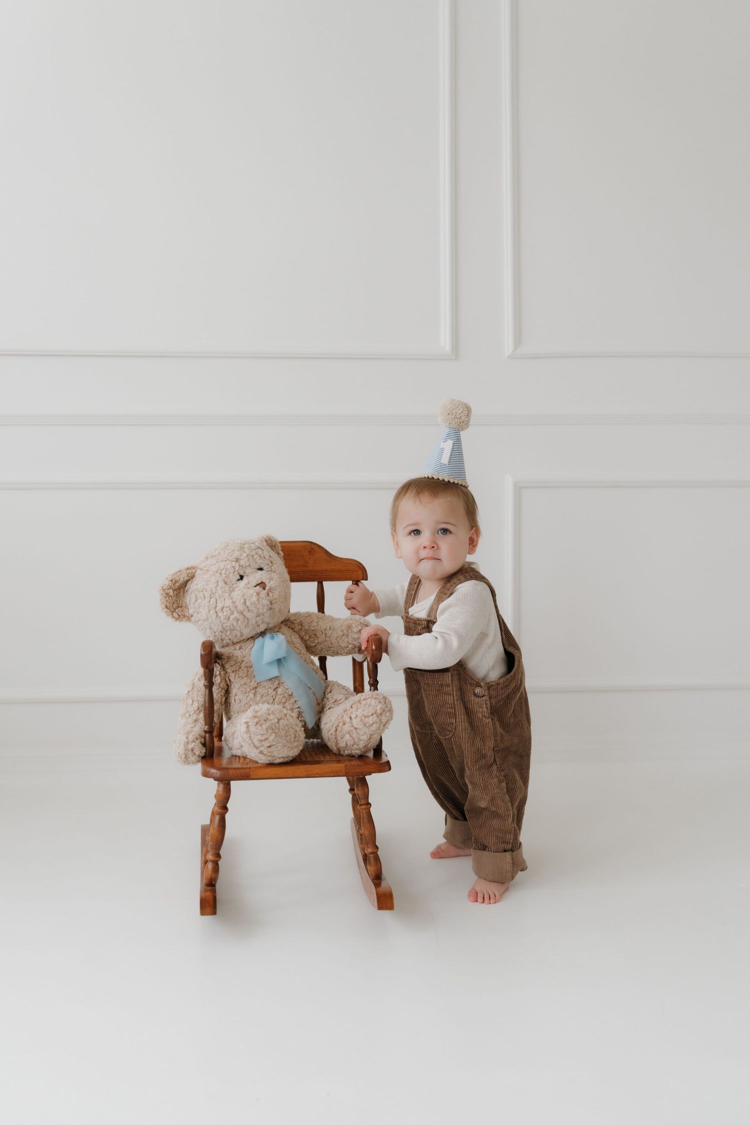 A small child in brown overalls stands next to a wooden chair with a large teddy bear in a minimalist white room.