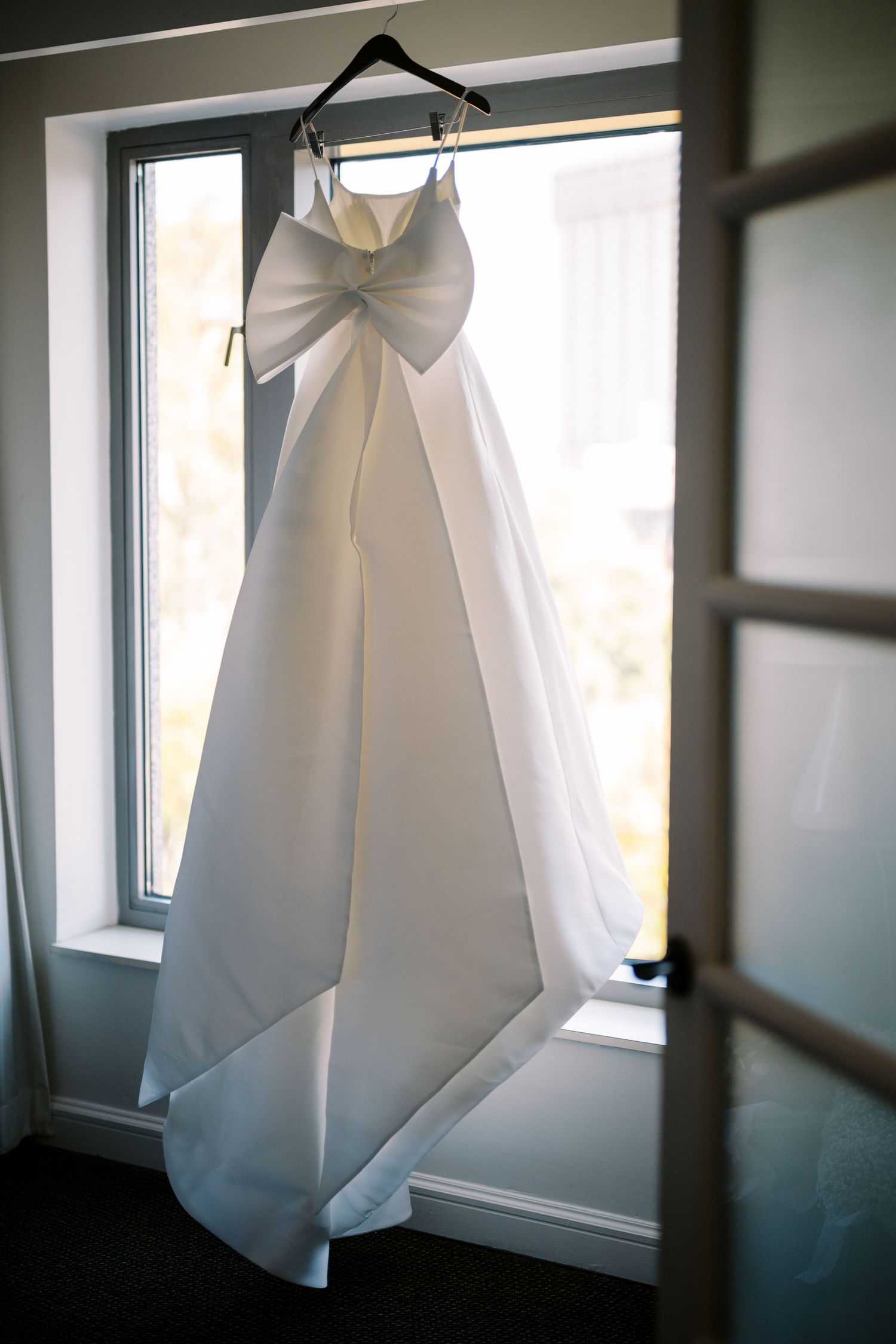 A white wedding dress with a large bow detail hangs elegantly by a sunlit window.