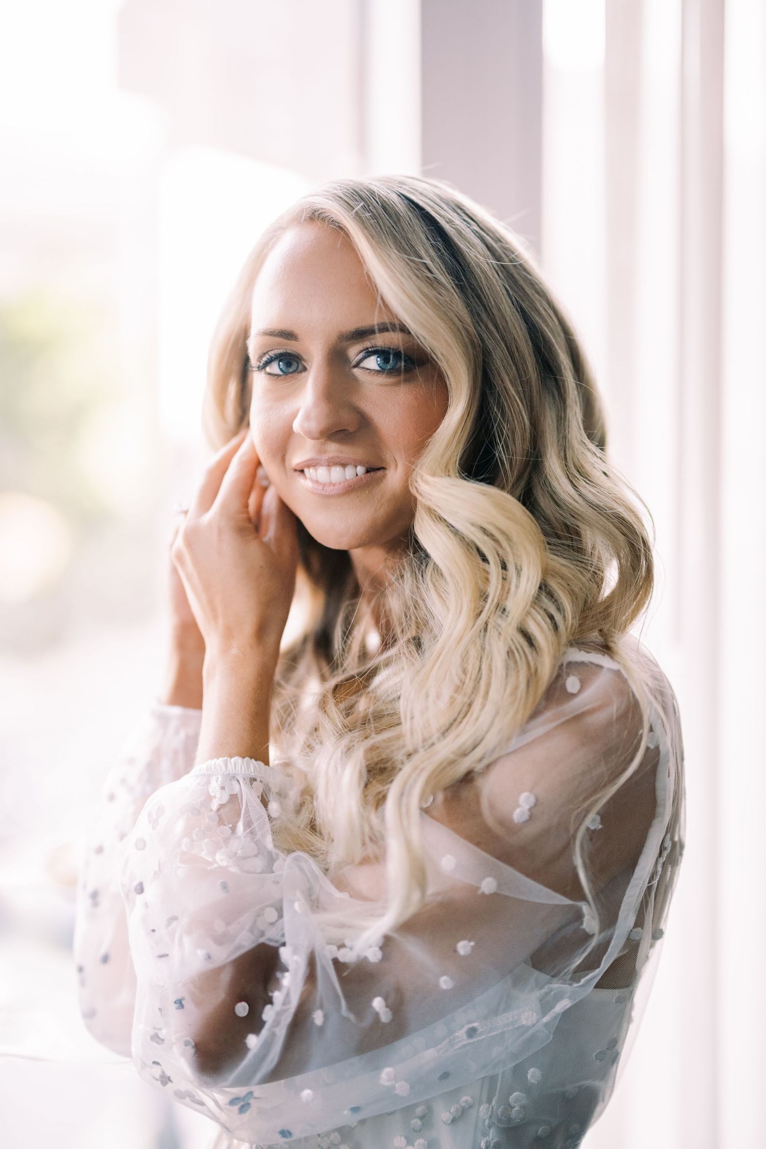 A bridal portrait showing a woman in a sheer white polka dot top with delicate blonde waves near a window.