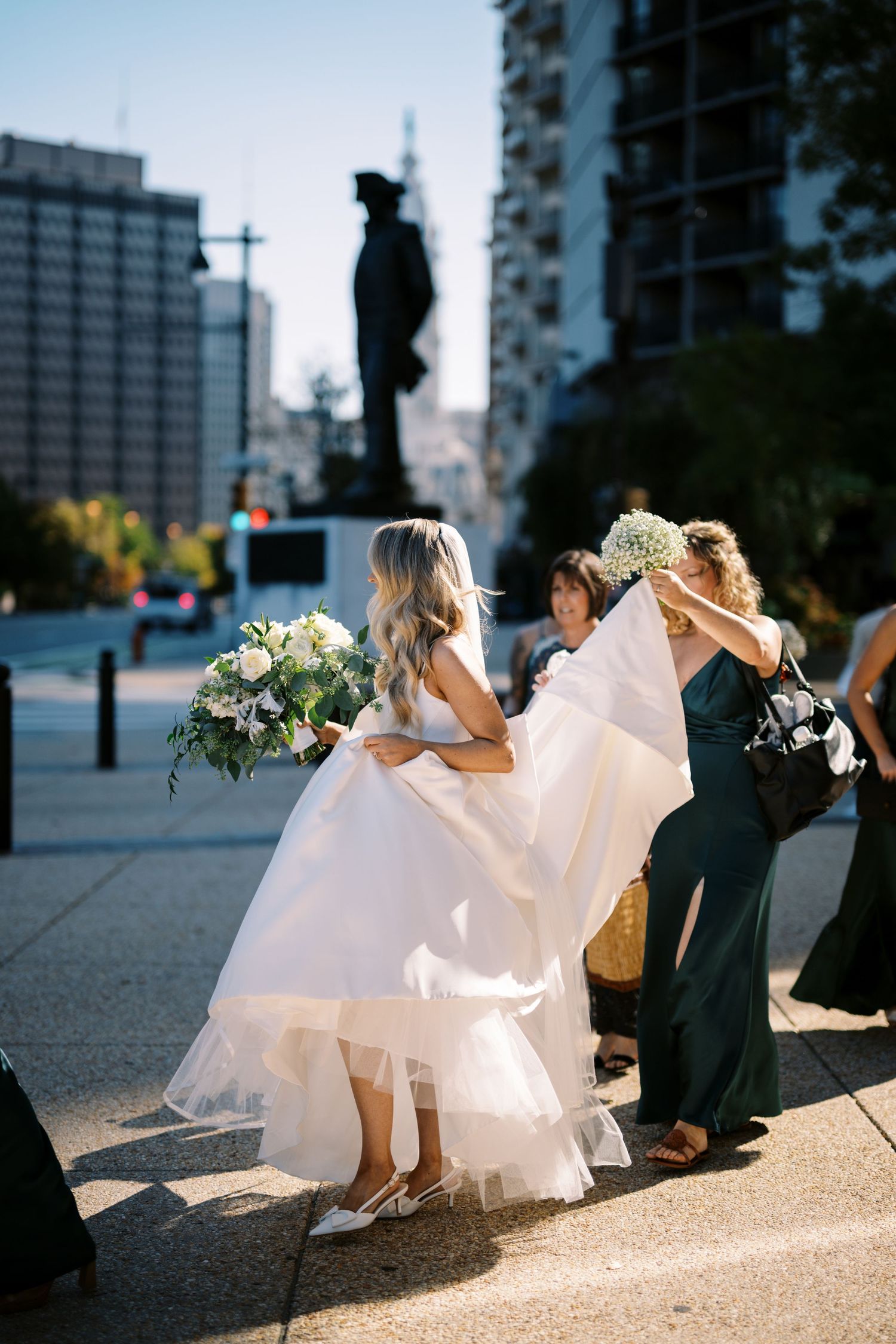 A wedding party stands on a city sidewalk at dusk as a person holds up a flowing white bridal gown train.