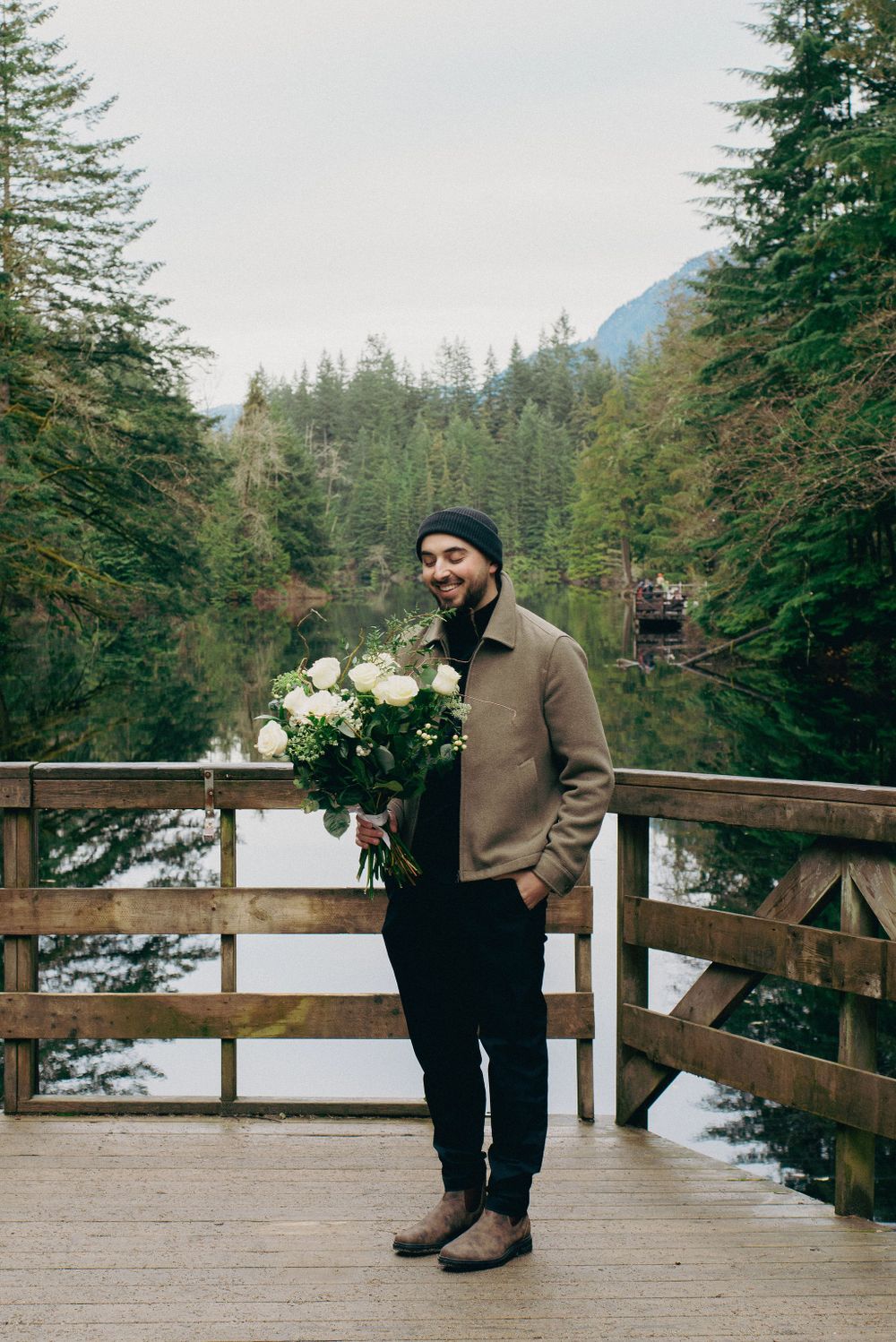 A person stands on a wooden deck overlooking a lake surrounded by evergreen trees.