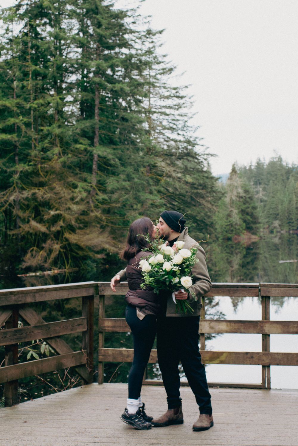 A romantic scene unfolds on a wooden deck overlooking a snow-dusted lake and forest.