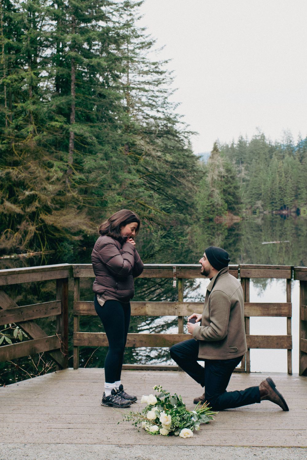 A romantic winter proposal unfolds on a scenic wooden deck overlooking a snowy forest landscape.