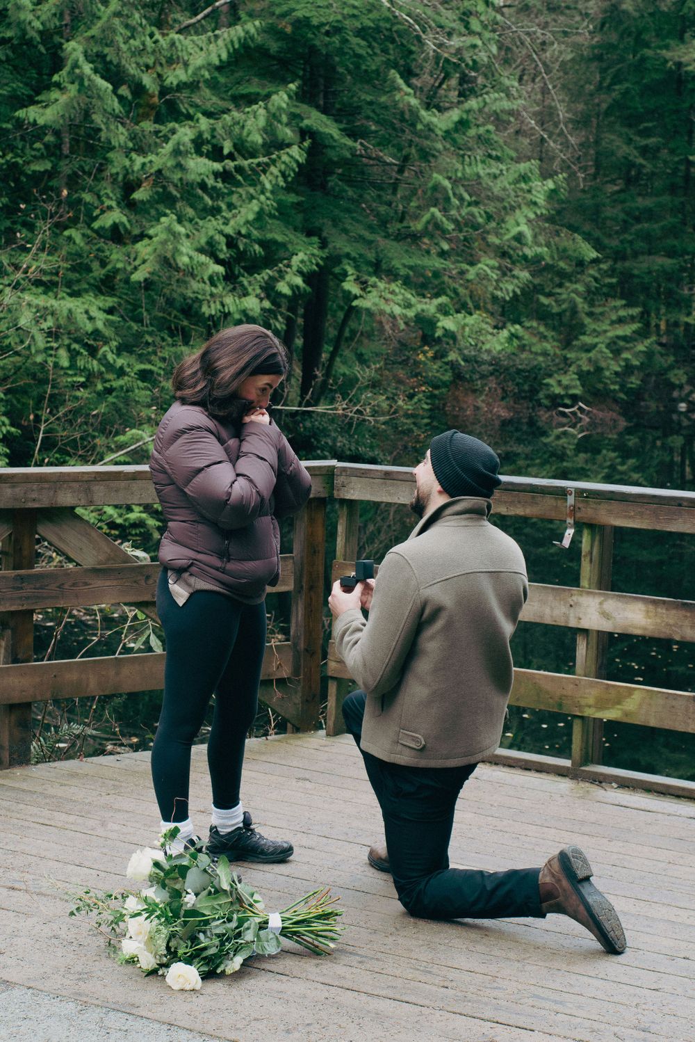 A couple shares a heartfelt moment during a proposal on a wooden viewing platform in nature.