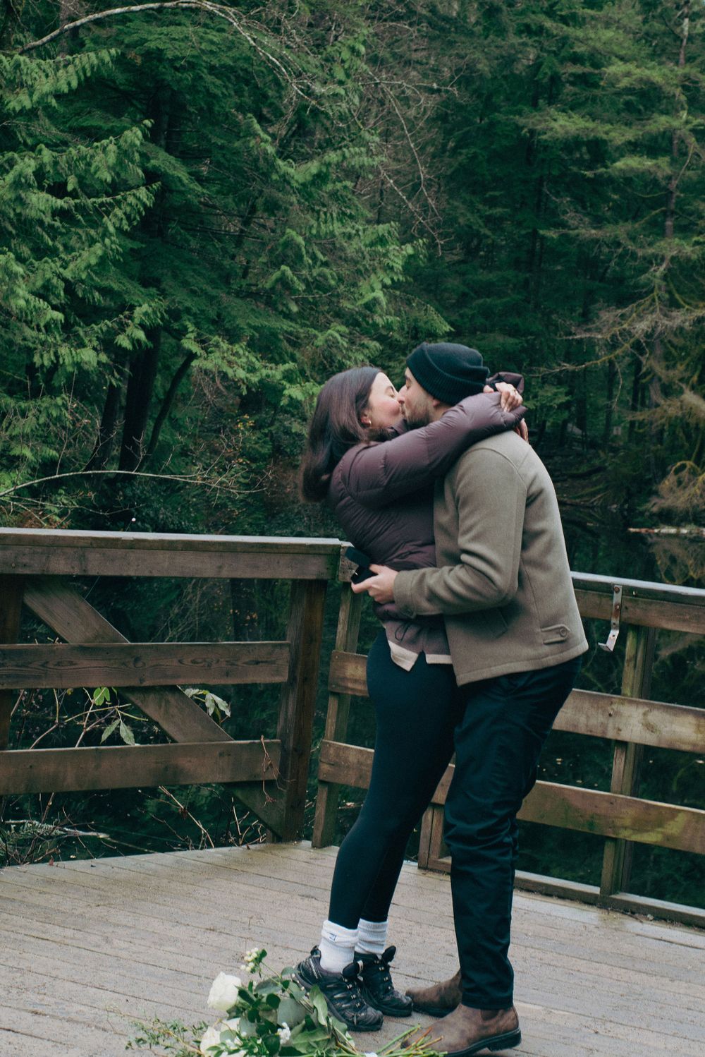 Two people embrace joyfully after a surprise proposal on a deck surrounded by evergreen trees.