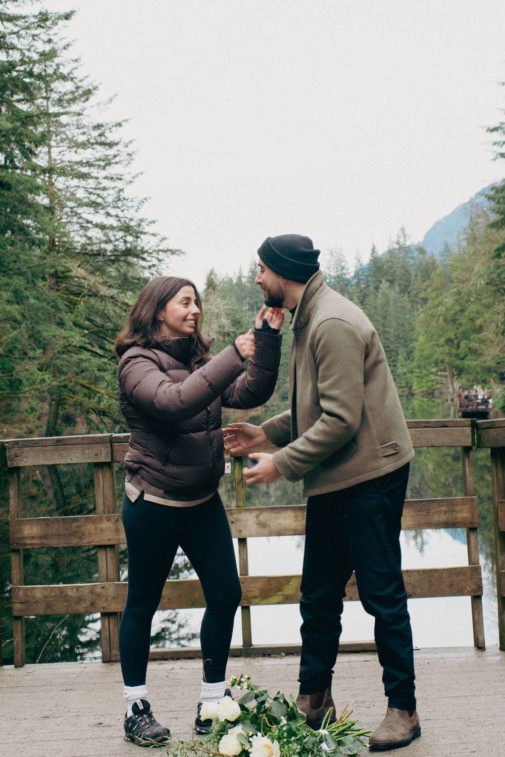 A couple shares excited reactions during a surprise proposal on a wooden deck in the wilderness.