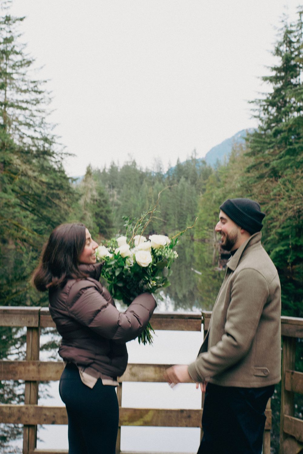Two people celebrate with joyful gestures during a romantic proposal on a forest viewing platform.
