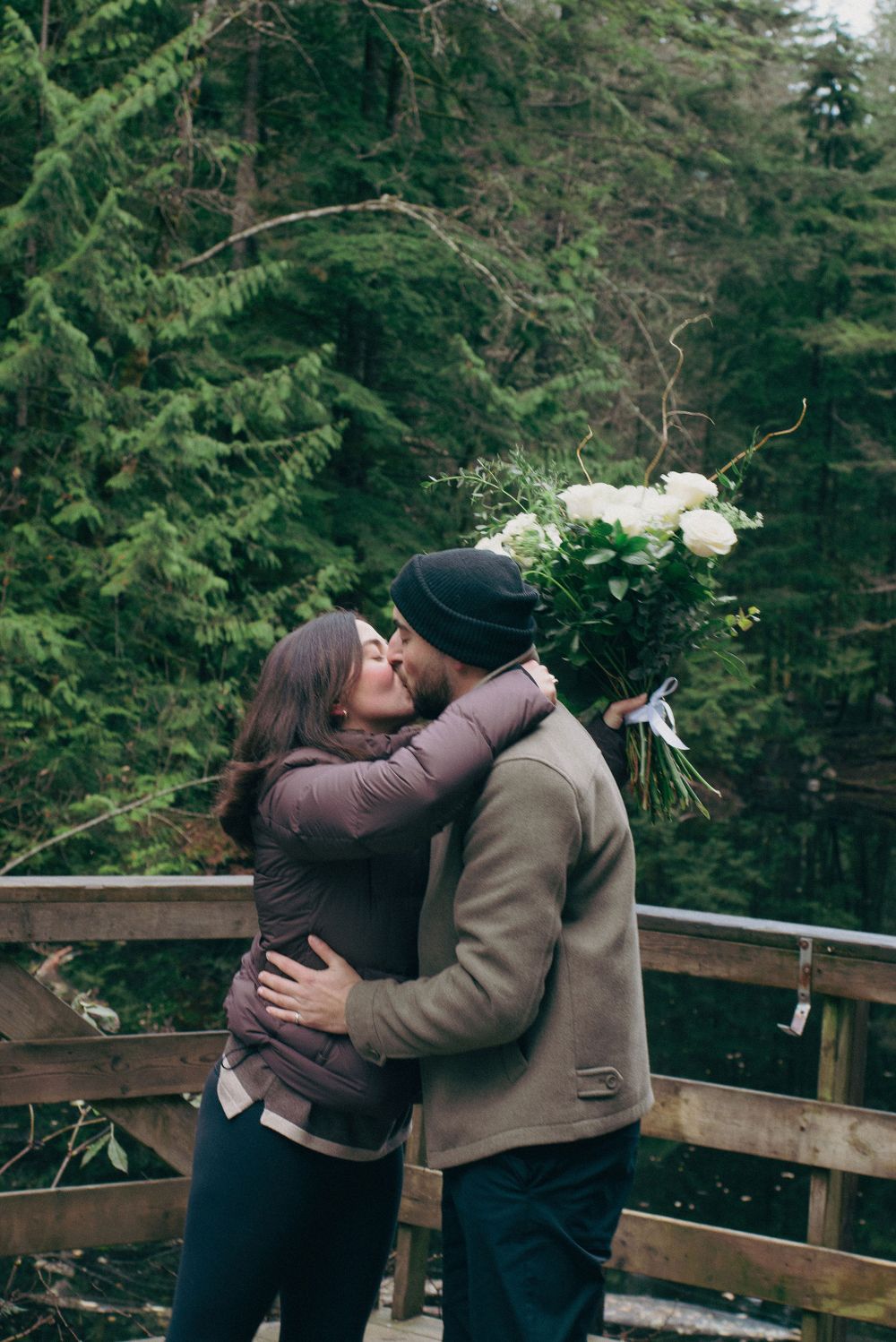 Two people share a romantic embrace surrounded by evergreen trees and snow-covered landscape.
