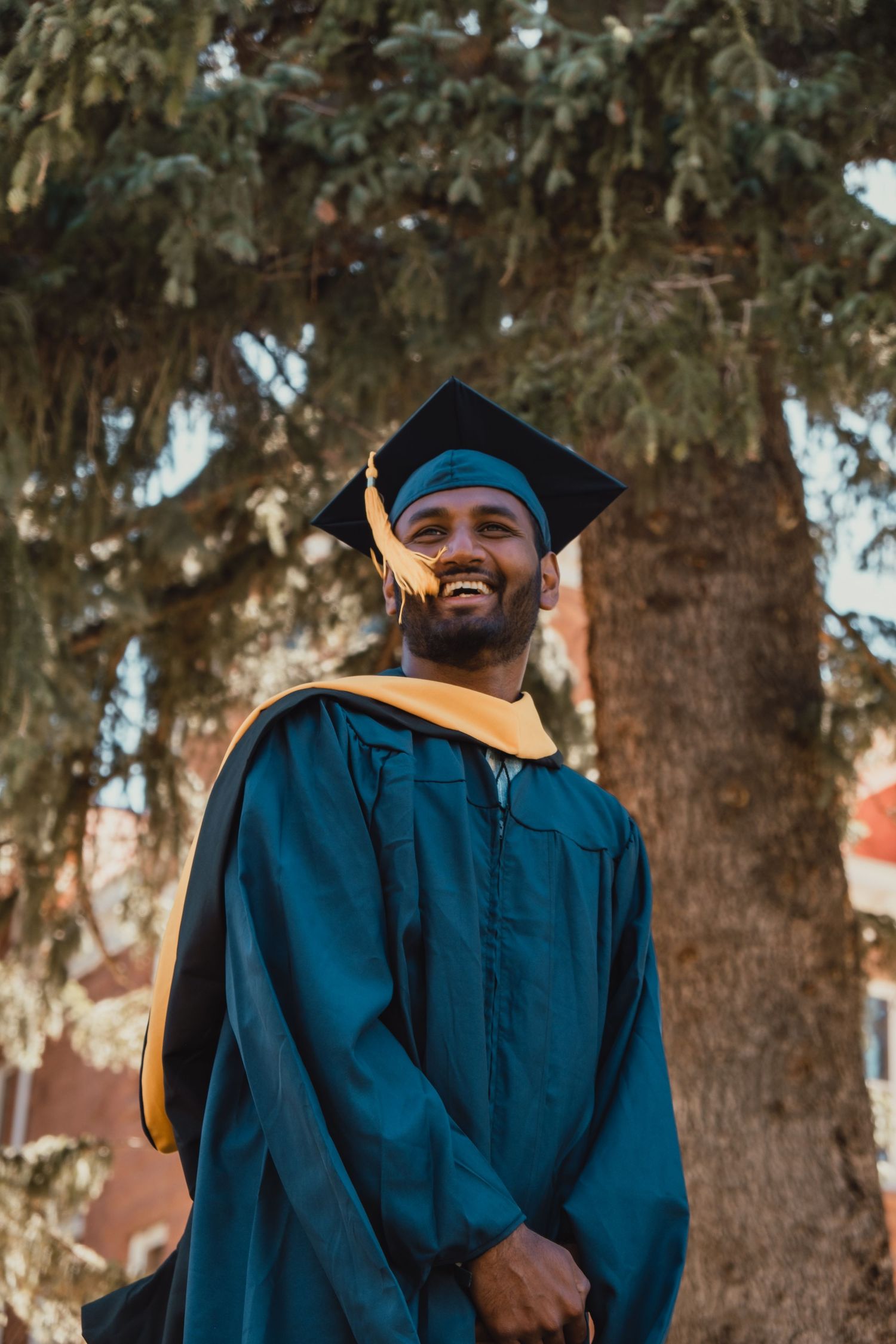 A graduate in a dark blue cap and gown with gold trim stands outdoors smiling in warm sunlight against a tree backdrop.