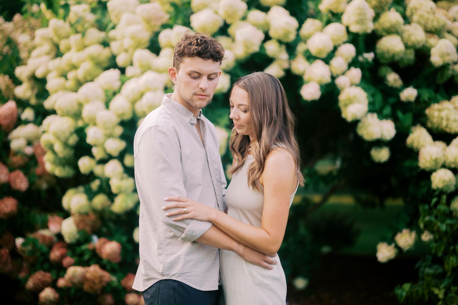 A couple shares an intimate moment in front of blooming white hydrangea bushes in a garden setting.