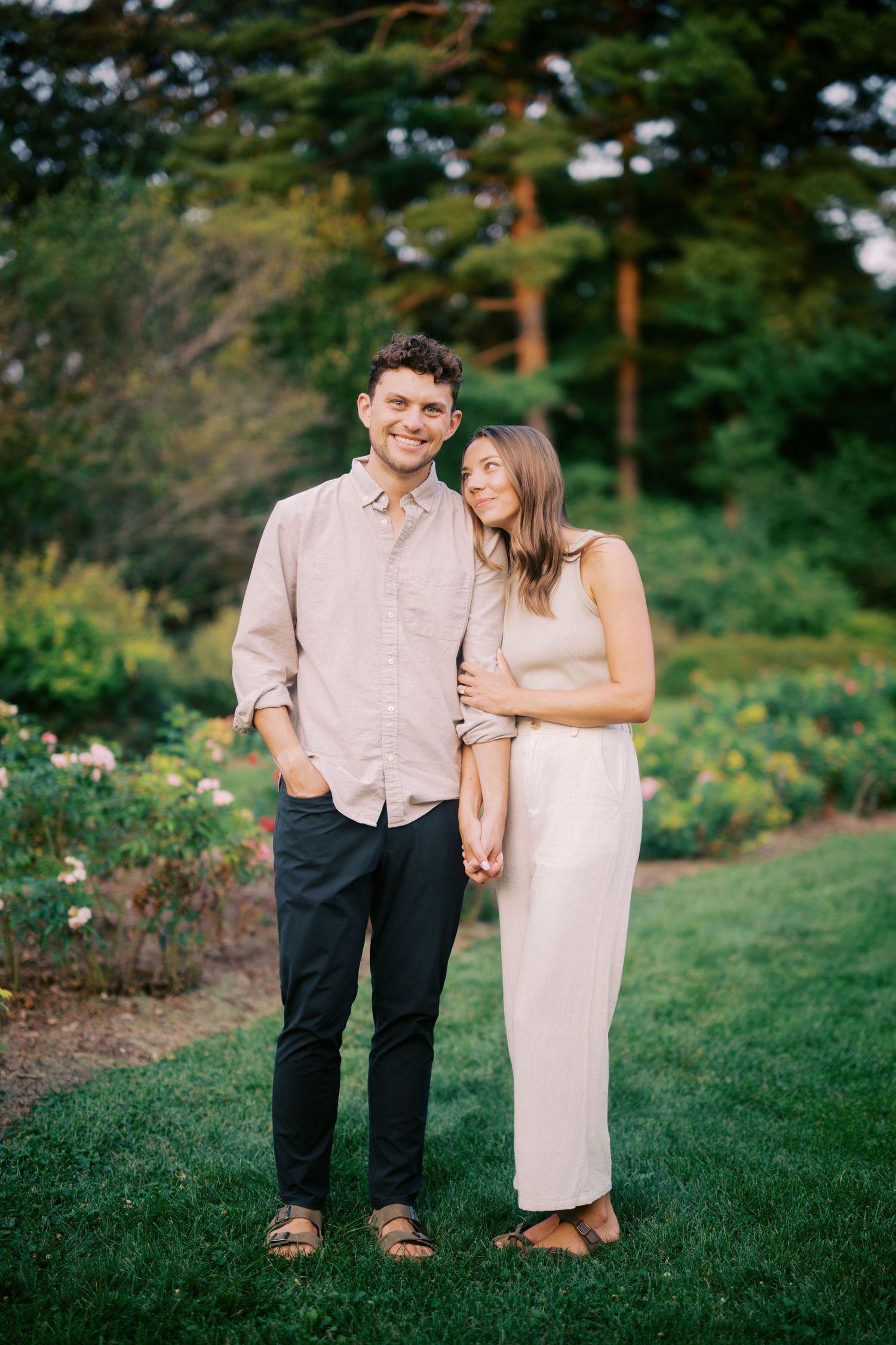 A couple in coordinated blush tones stands together on a manicured lawn with lush greenery in the background.