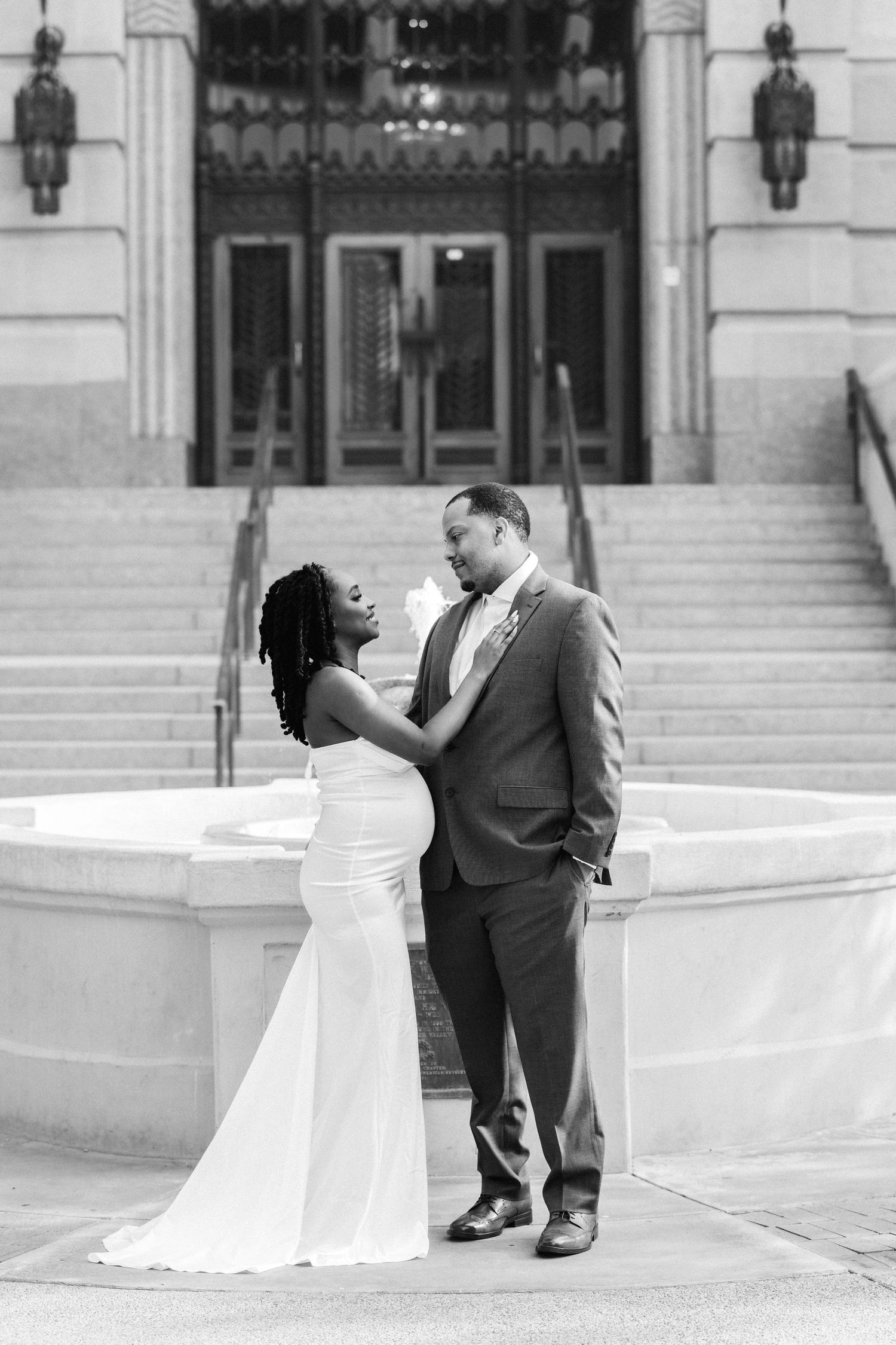 A loving couple in formal wedding attire share an intimate moment on elegant stone steps of a grand building entrance.