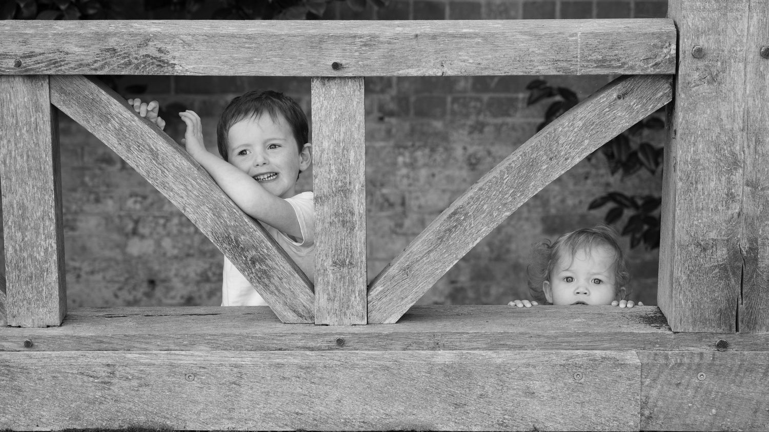 Two young children peek through the gaps of a rustic wooden fence structure in a black and white photo.